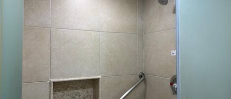 A shower area with beige tiles, a handheld showerhead, a grab bar, and a shelf with toiletries.