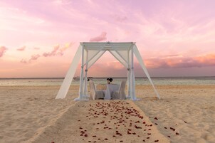 Decoración para boda en la playa con una marquesina y una mesa, rodeado de arena y pétalos de rosa.