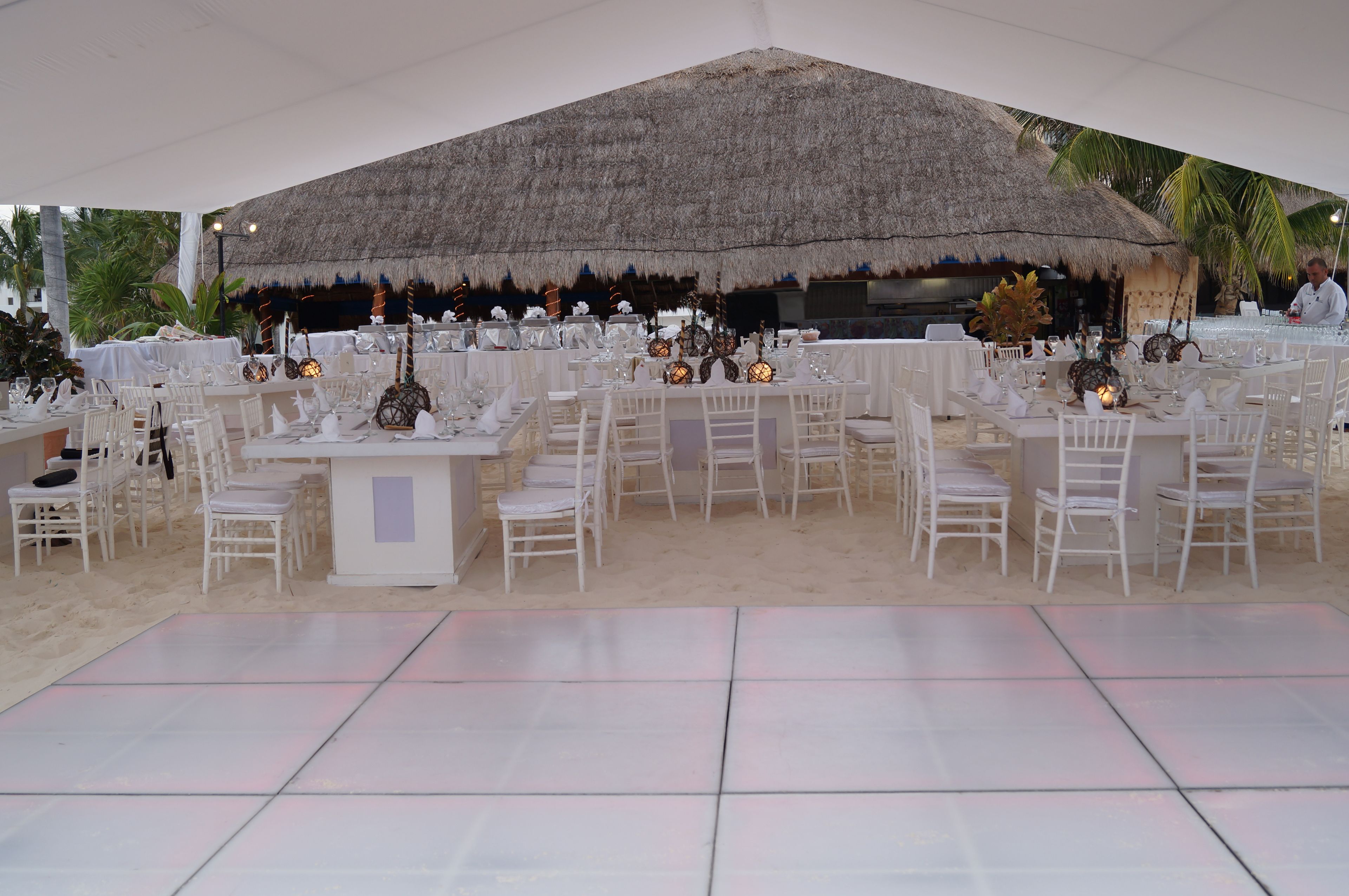 A beachside event space with thatched-roof tables and chairs, arranged on a tiled floor.