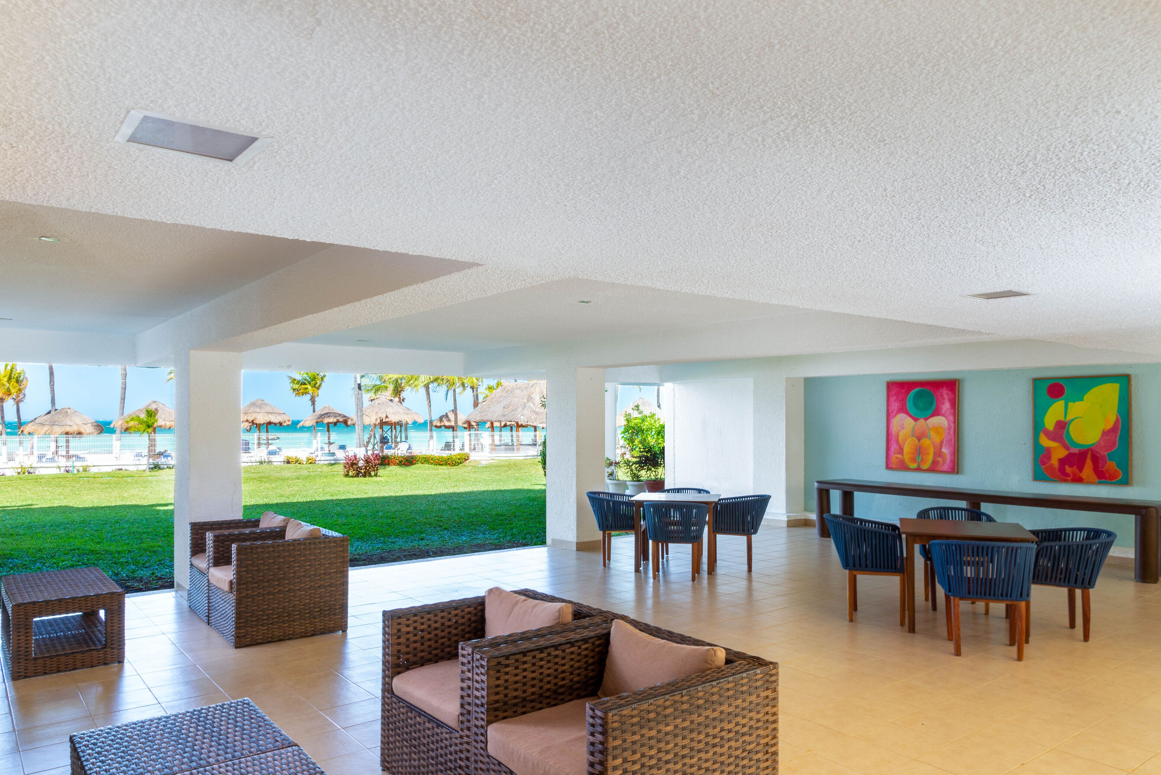 A spacious indoor area with wicker furniture, a dining table, and chairs. There are paintings on the wall and a view of a pool and palm trees outside.
