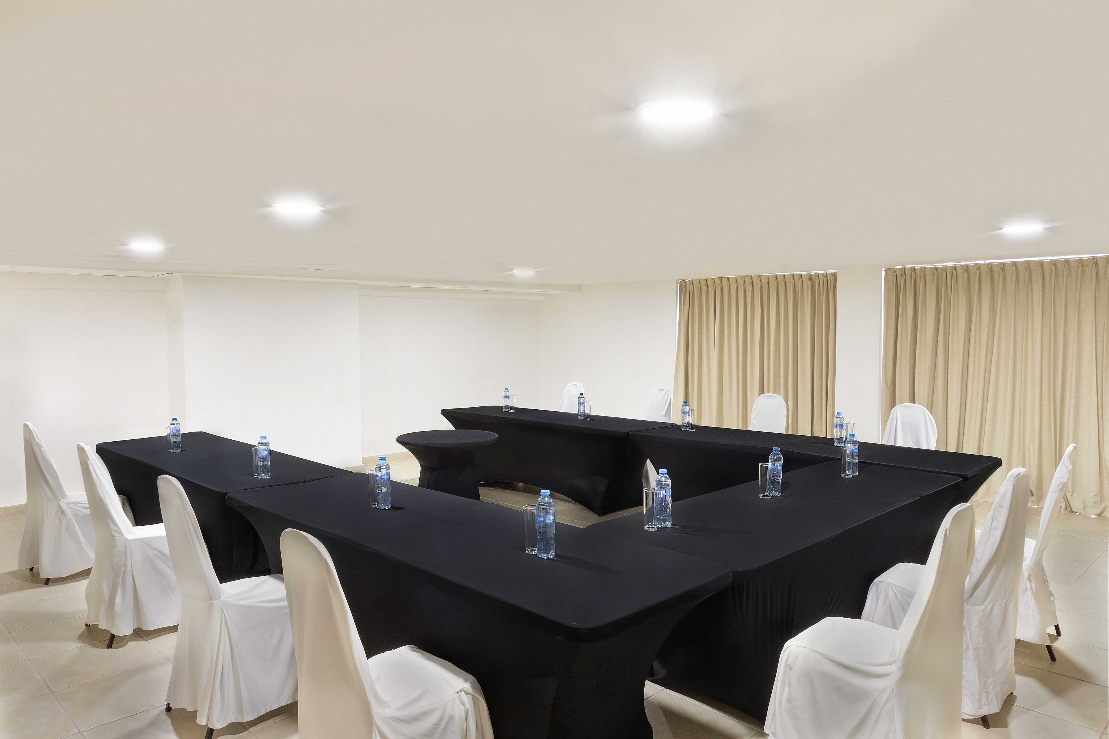Une salle de conférence avec une longue table, des chaises blanches et des bouteilles d’eau posées sur la table.