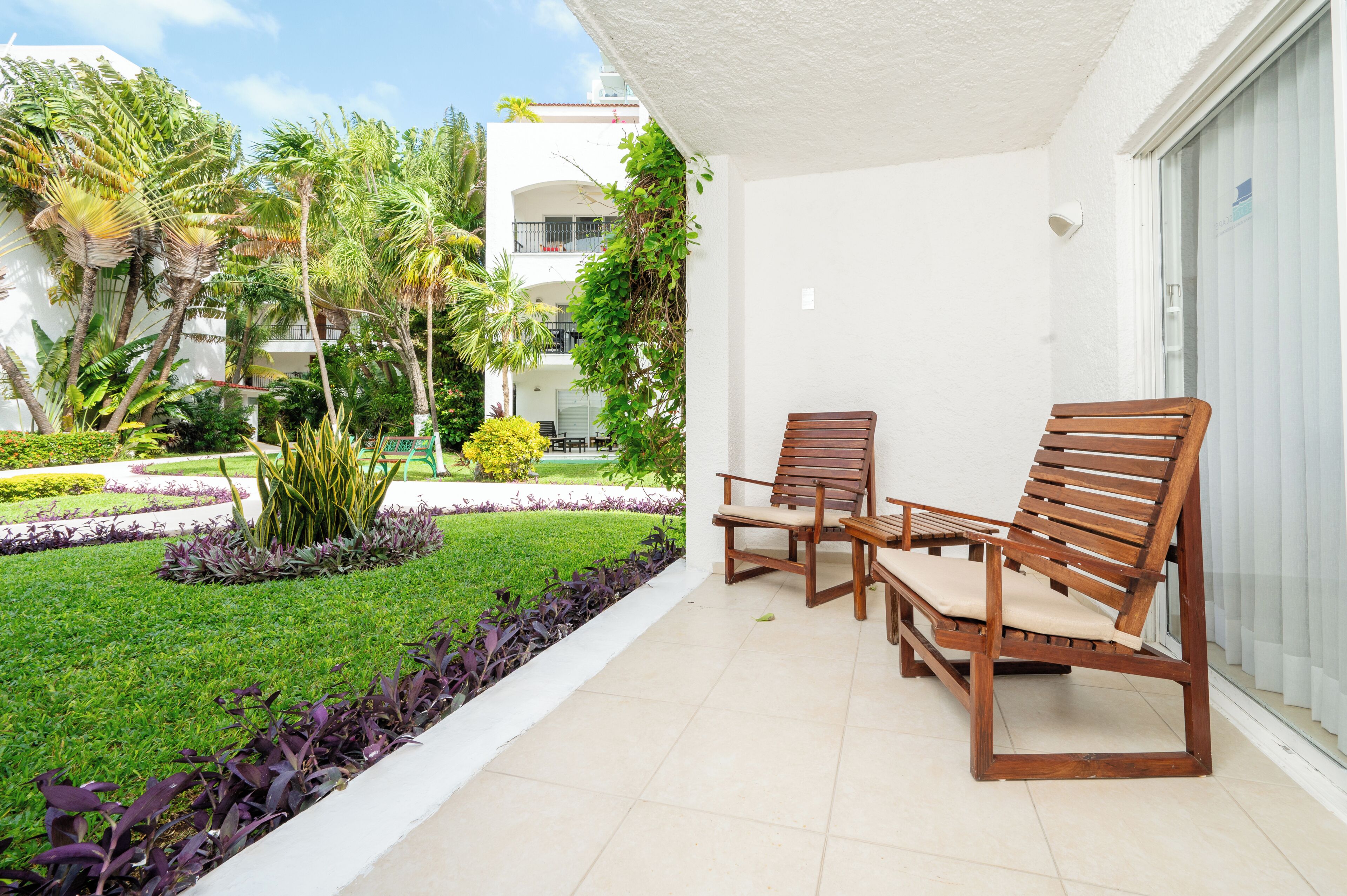 A balcony with two wooden chairs and a small table, surrounded by a well-maintained garden with various plants and a white building in the background.