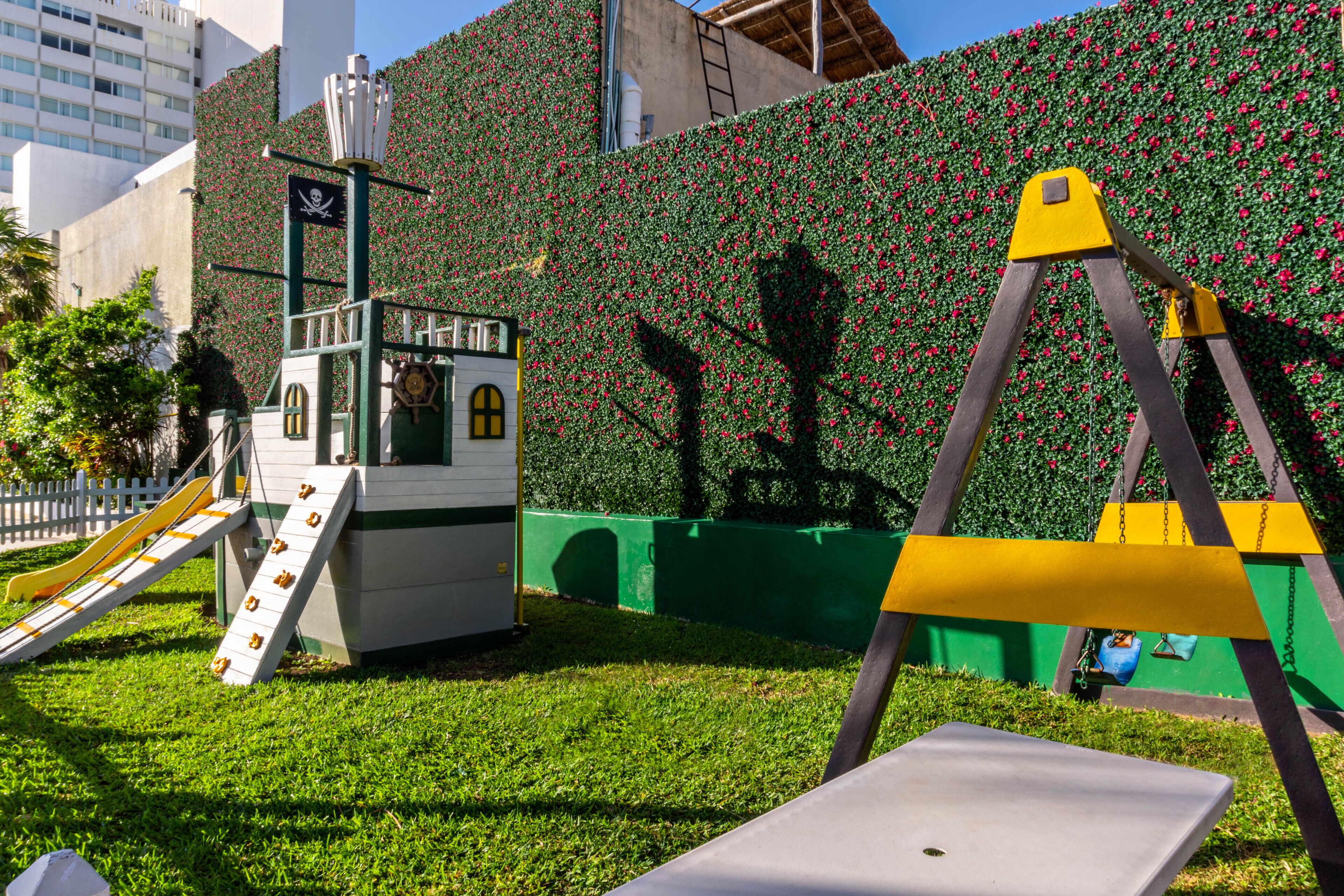 Un parque infantil con tobogán y escaleras de acceso, un columpio y un pequeño edificio con una bandera pirata.