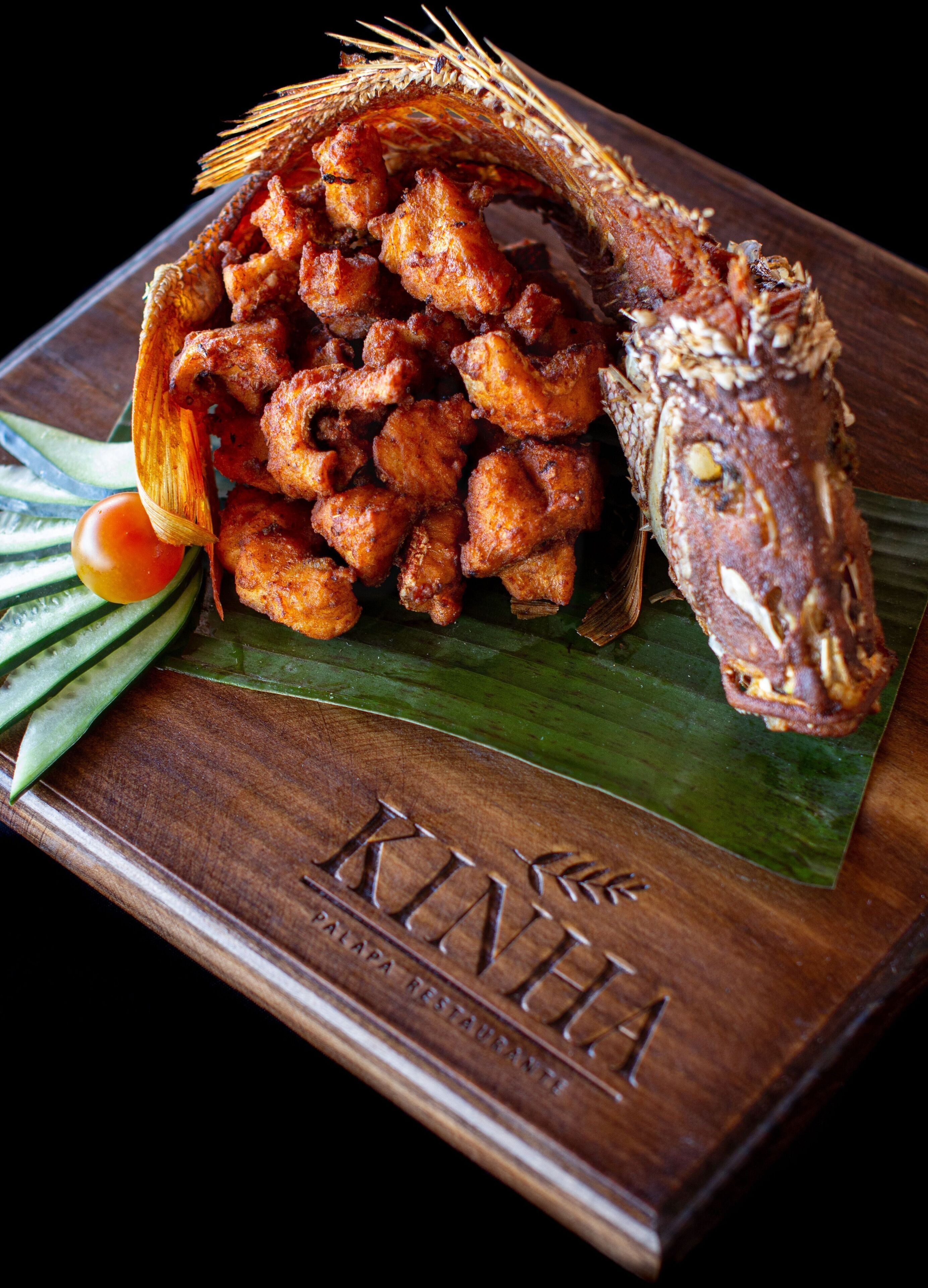 A wooden serving board with fried fish and vegetables, branded with "KINHA RESTAURANT".