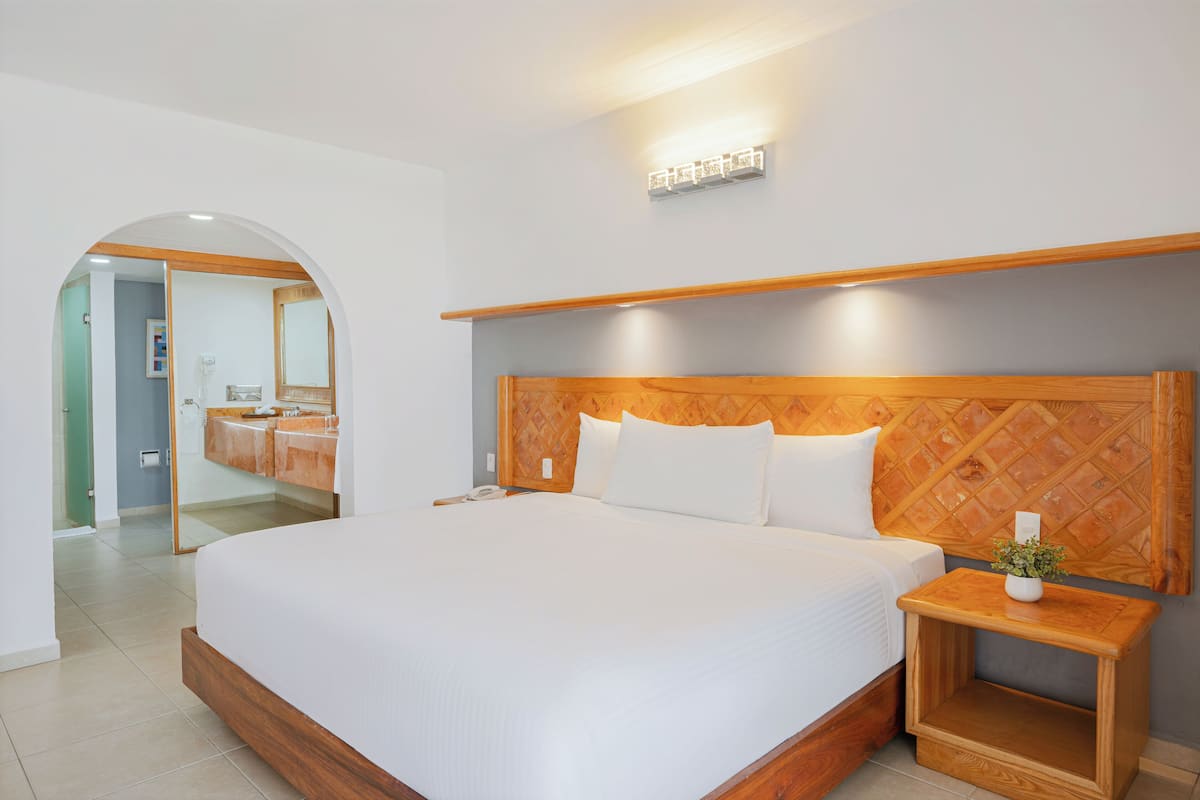 A modern hotel room with a large bed, wooden headboard, bedside tables, and a bathroom visible through an arched doorway.