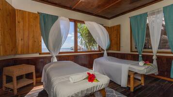 Two single beds with white linens and a small bench, overlooking the sea and palm trees.