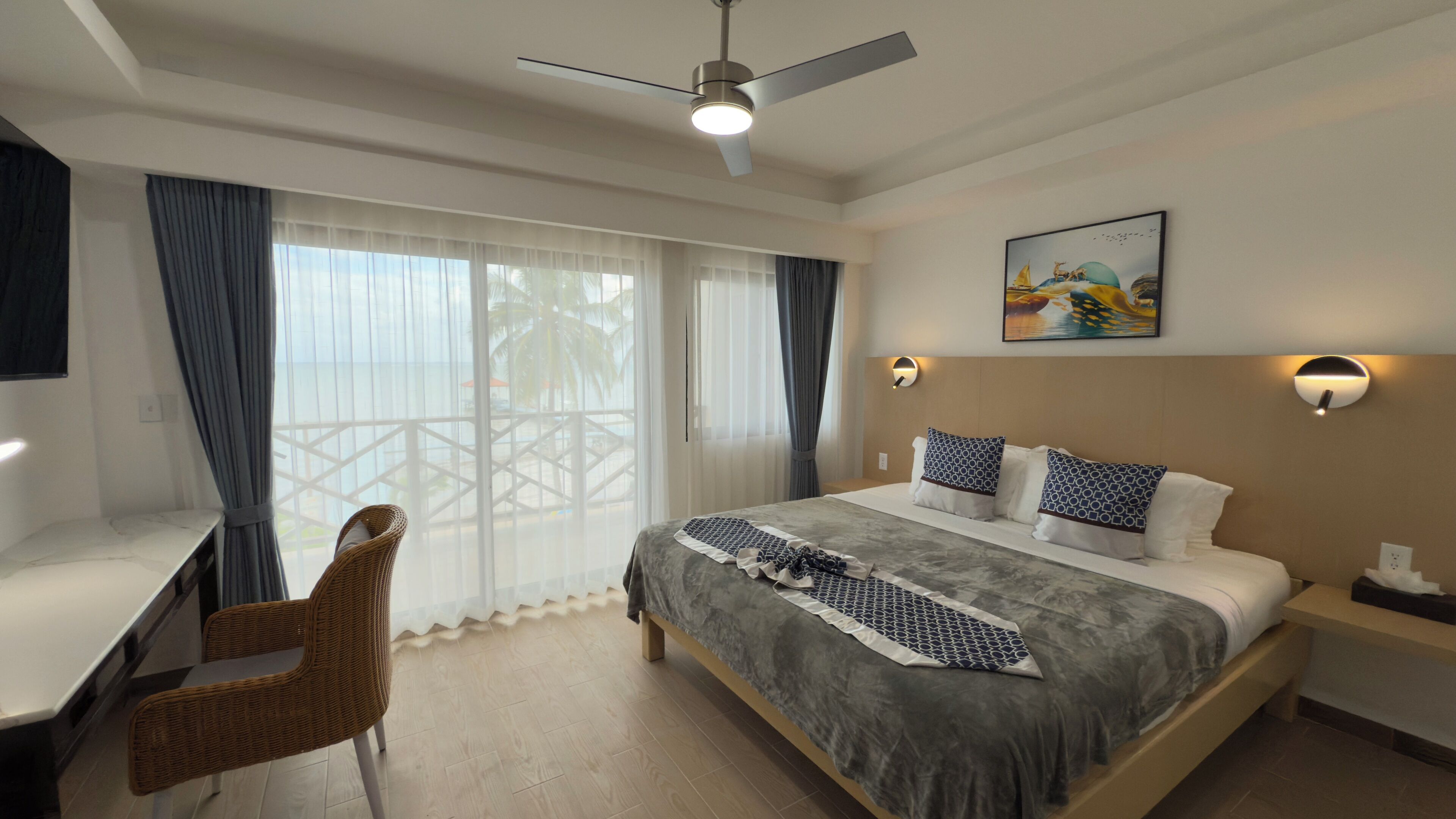 Une chambre d’hôtel équipée d’un lit, d’un bureau, d’une chaise, d’un ventilateur de plafond et d’une grande fenêtre avec des rideaux.