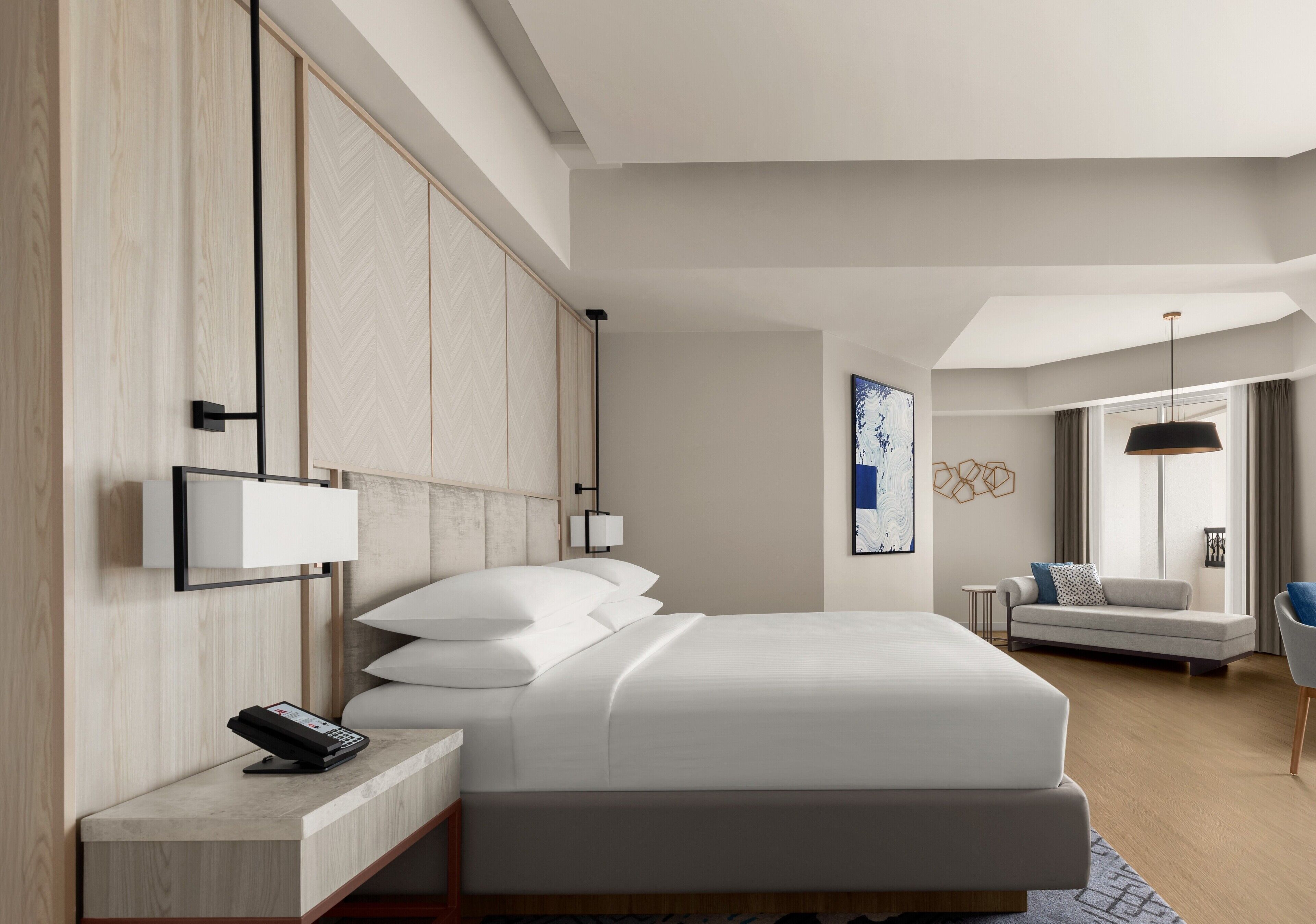 A modern hotel room with a large bed, bedside tables, a desk, a sofa, and a large window.