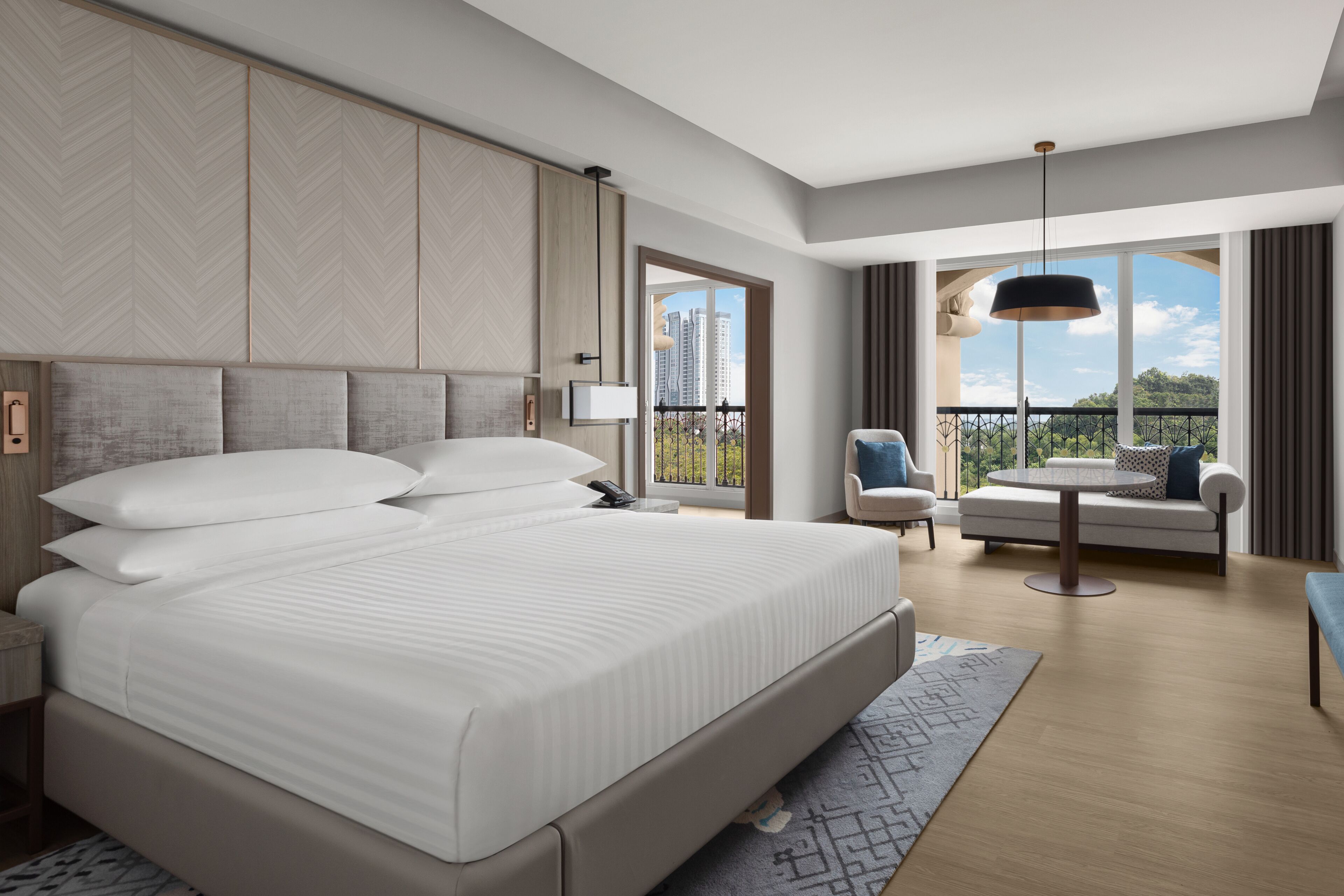 A modern hotel room with a large bed, a sofa, a chair, and a balcony with a city view.