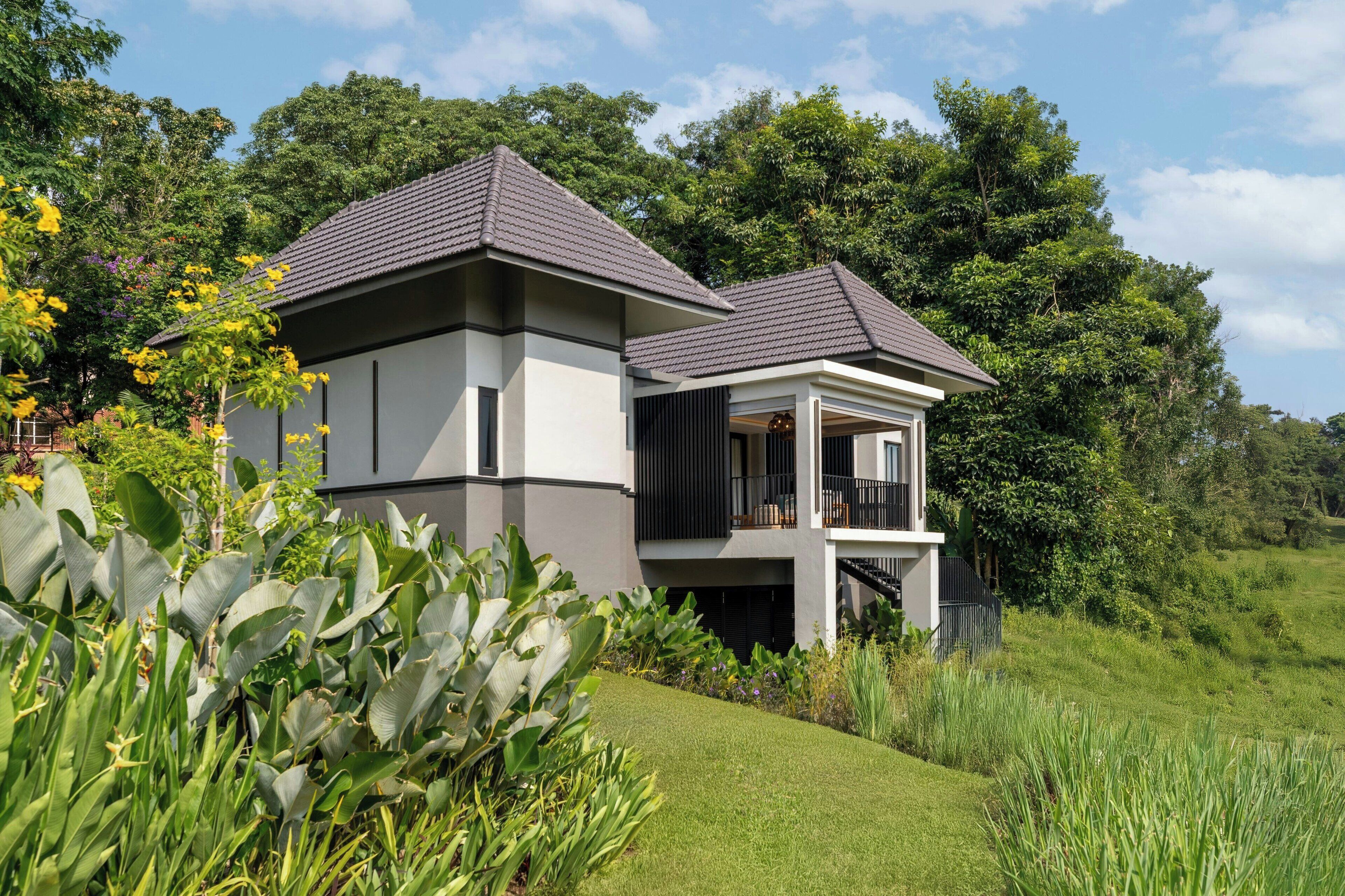 A modern two-story house with a balcony, surrounded by lush greenery and a well-maintained lawn.