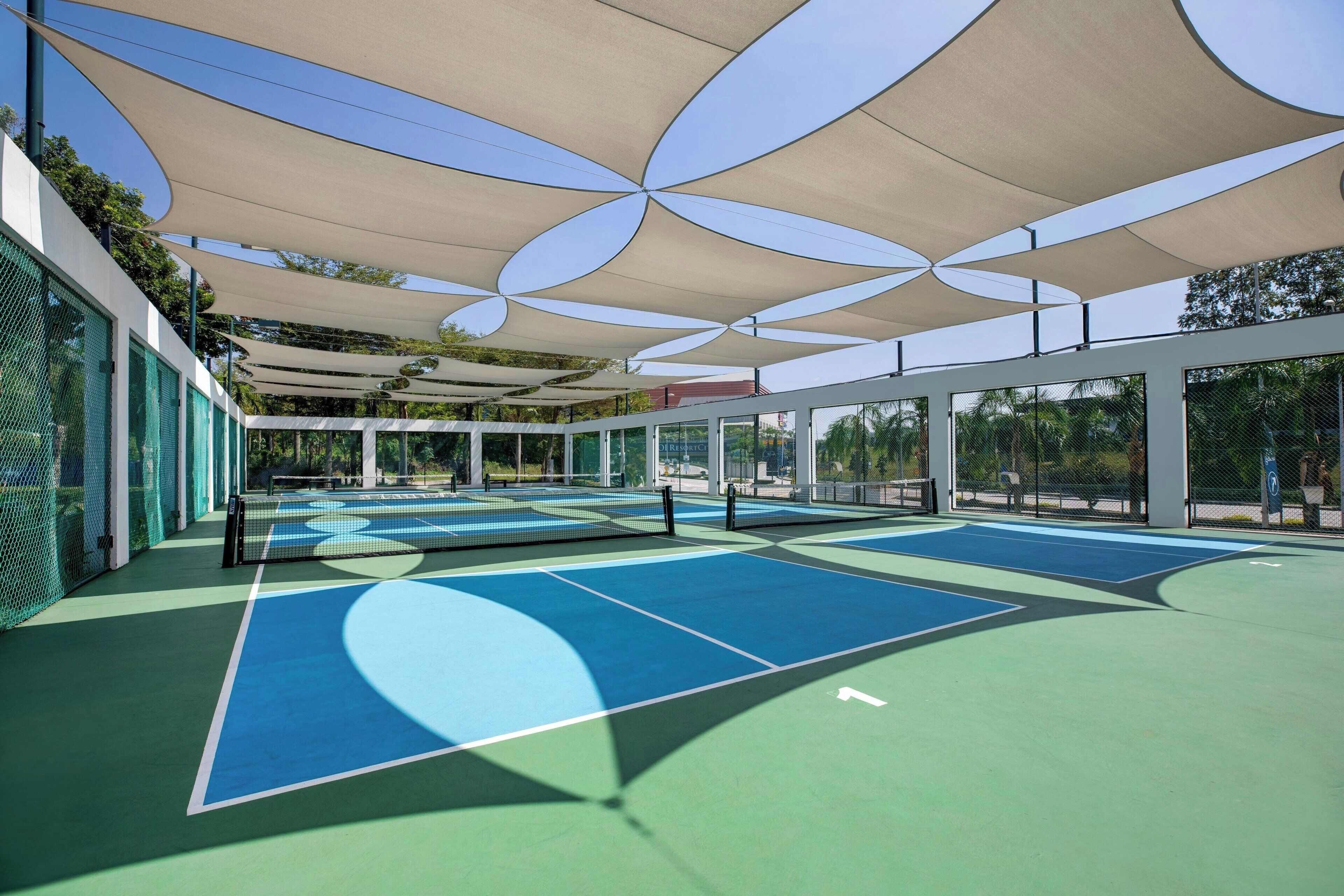 An outdoor tennis court with multiple courts, covered by large shade structures, and surrounded by glass walls.