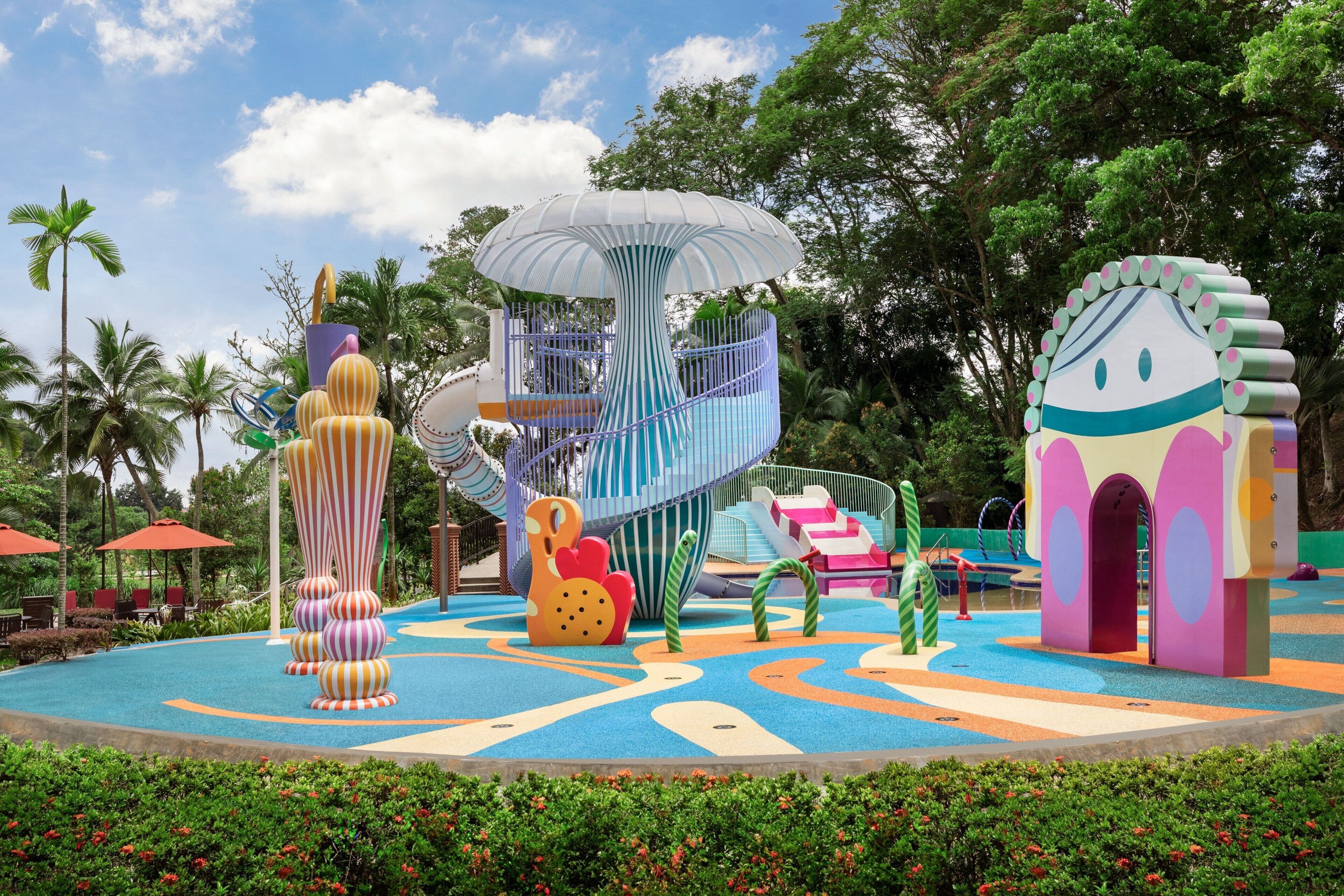 A colorful playground with various play structures, surrounded by lush greenery and palm trees.