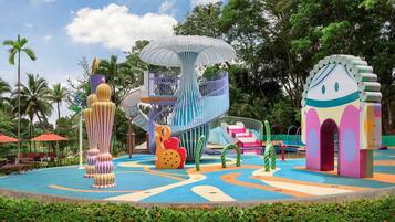 A colorful playground with various play structures, surrounded by lush greenery and palm trees.