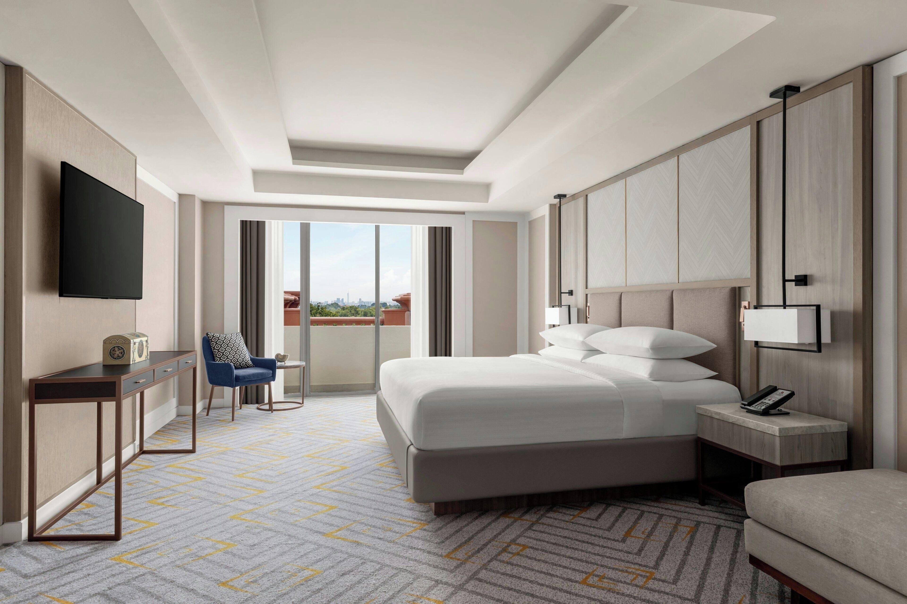 A modern hotel room with a large bed, a desk, a chair, a TV, and a balcony with a city view.