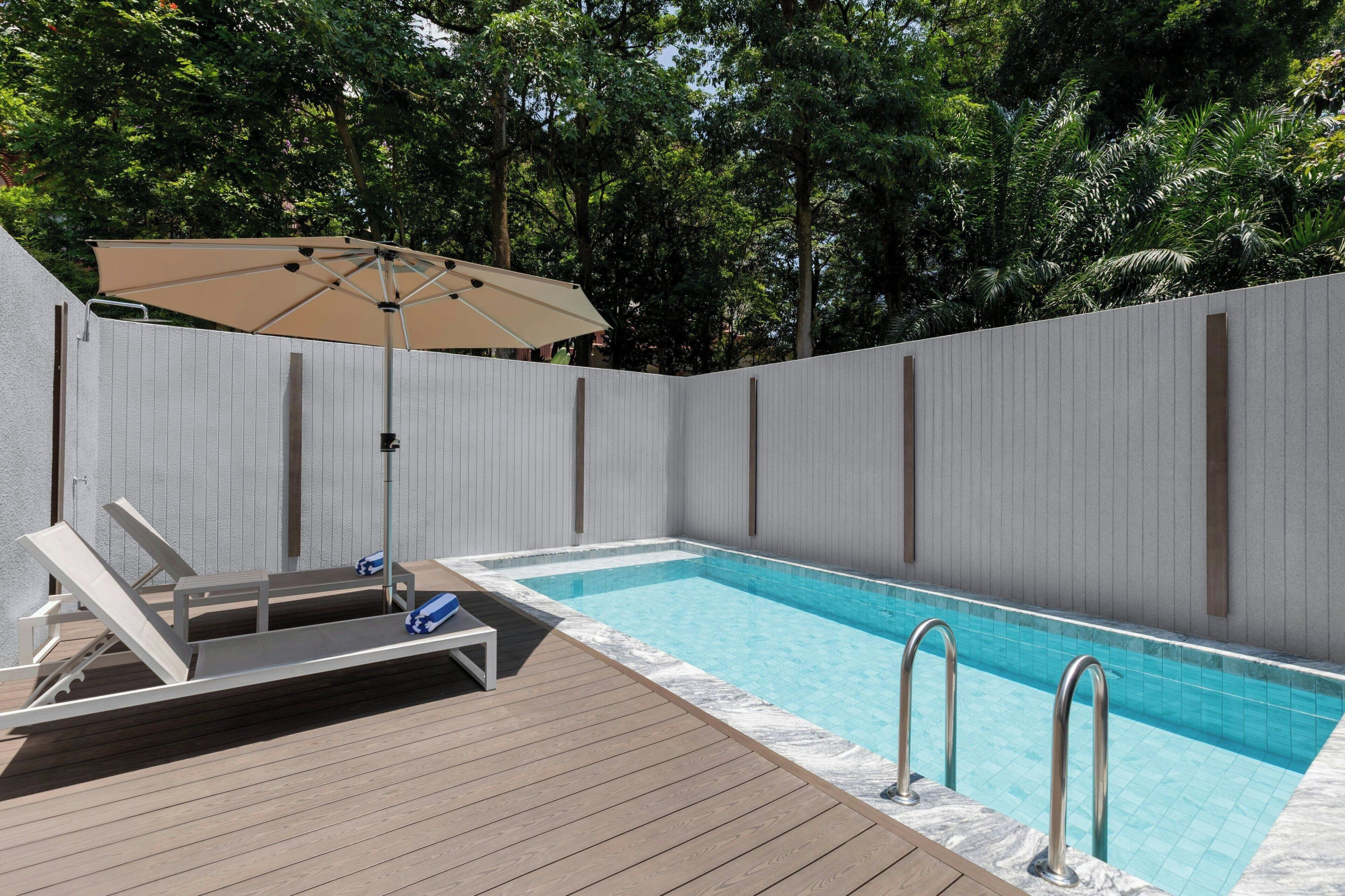 A rectangular swimming pool with a wooden deck, a lounge chair, and a beige umbrella.