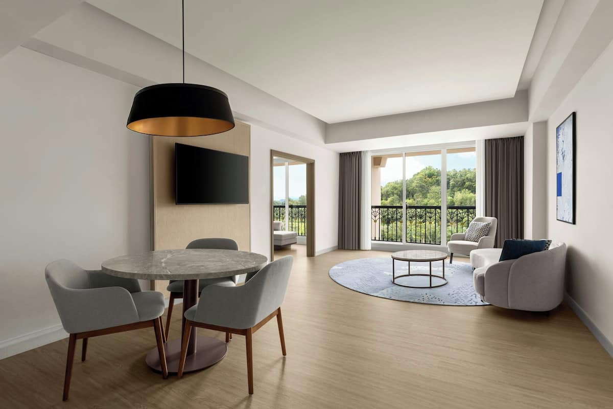 Una sala de estar moderna con una mesa de comedor redonda, sillas grises, un sofá y un balcón con vistas a zonas verdes.