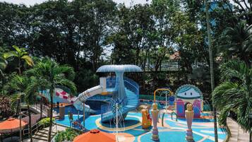 Parque acuático con tobogán, estructuras de juego y zona de piscina rodeada de palmeras y follaje.
