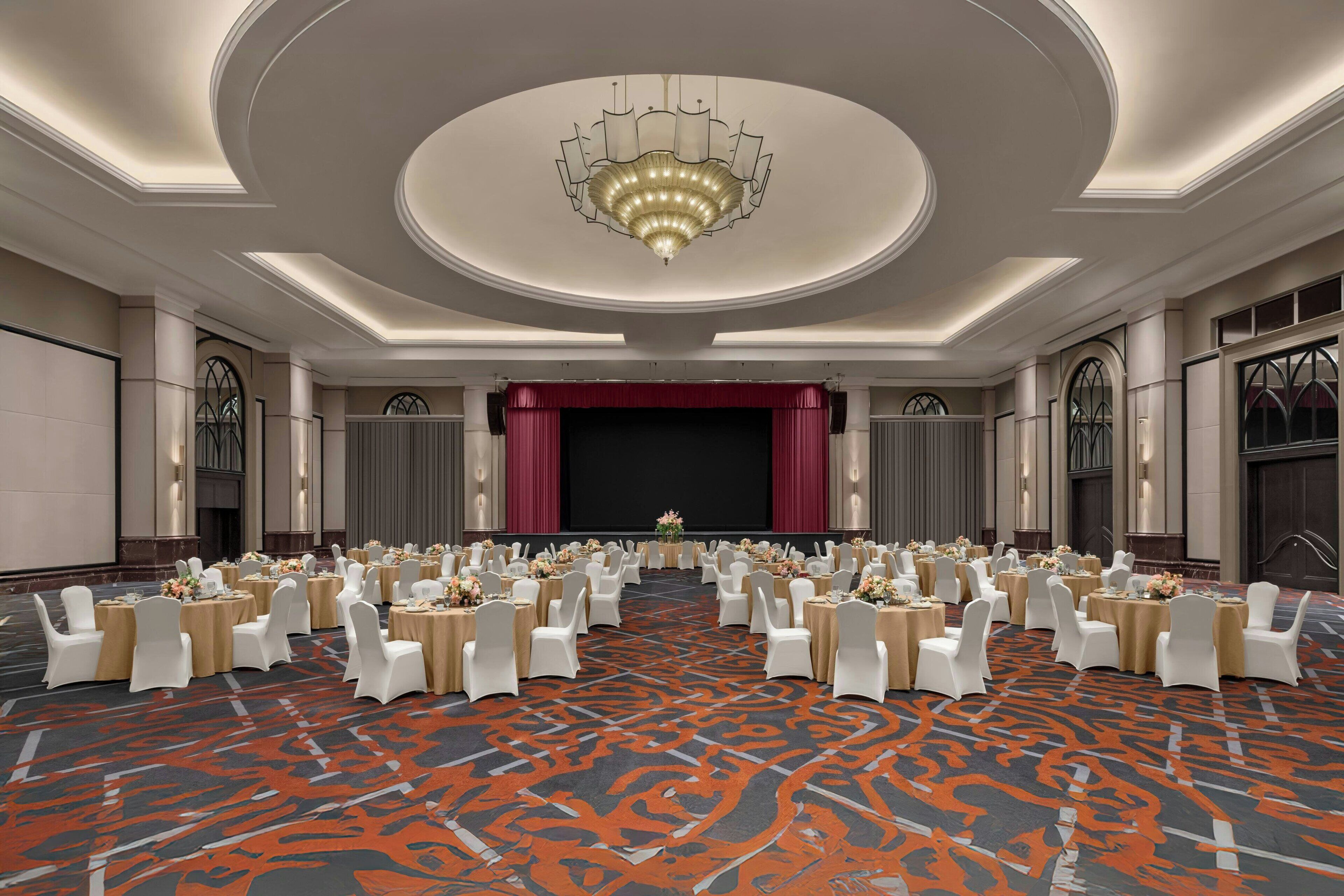 A large ballroom with a central stage, rows of round tables with white chairs, and a patterned carpet.