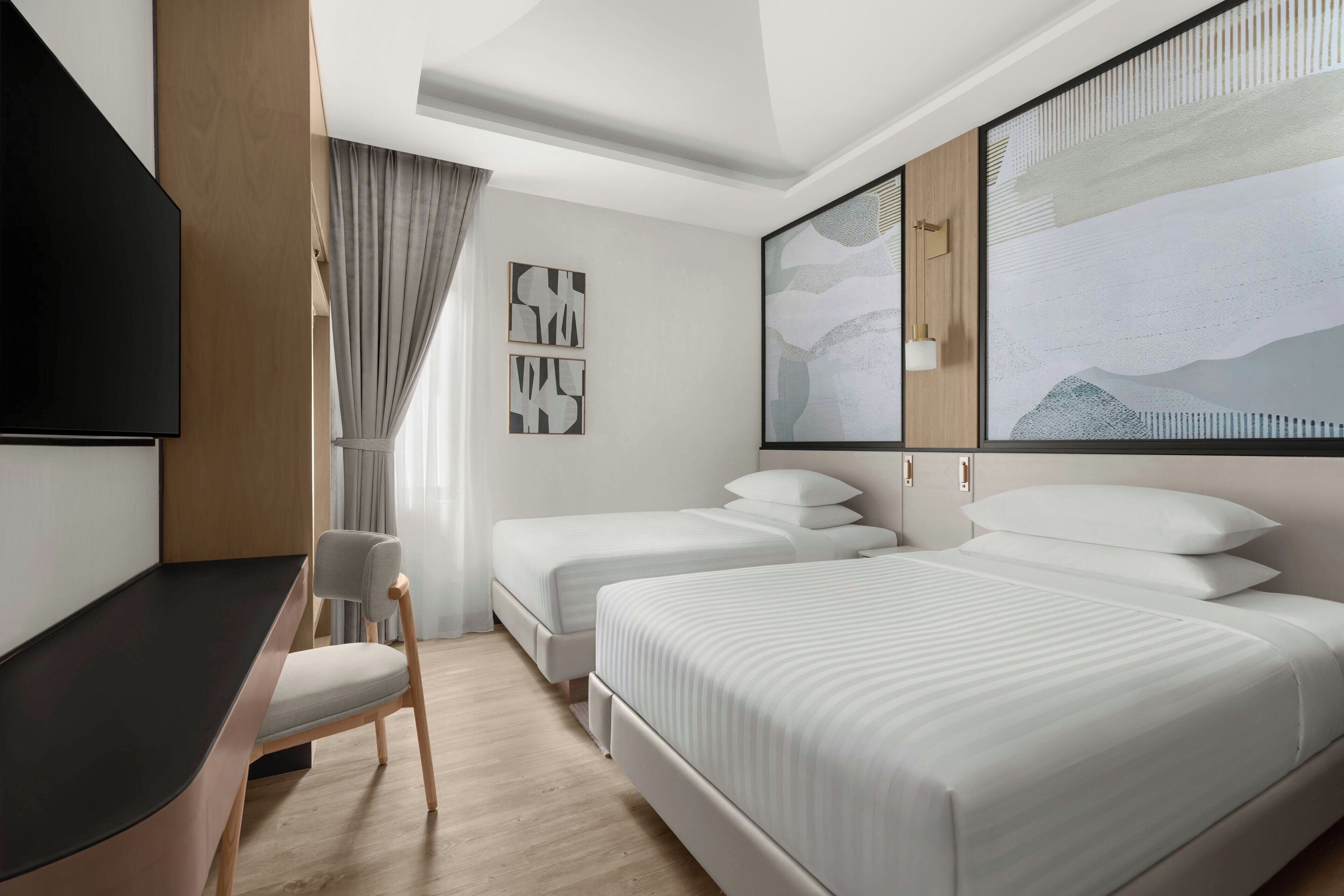 A hotel room with a large bed, two smaller beds, a desk, and a chair. There are abstract wall art pieces and a large window with curtains.
