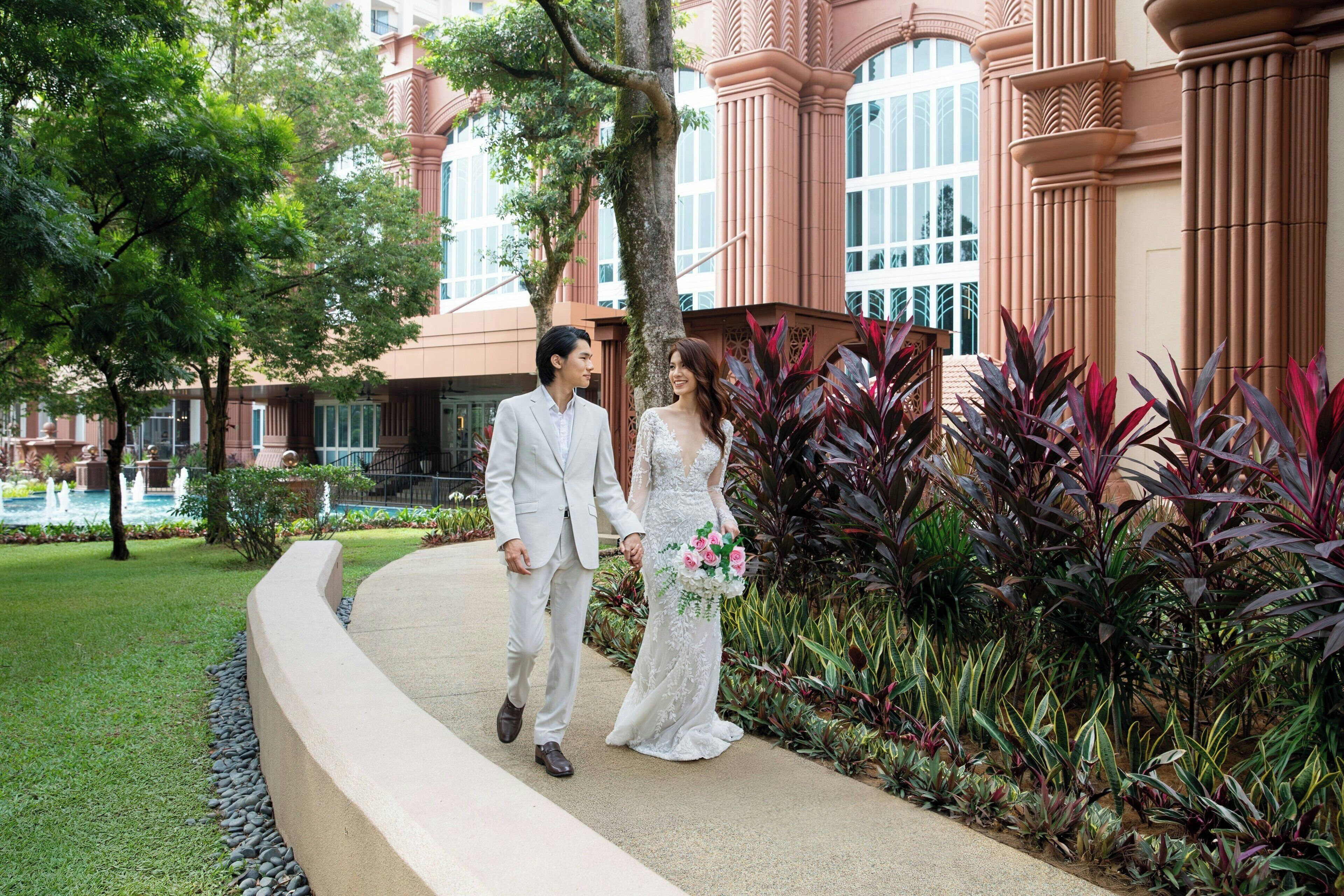 A couple walking on a pathway surrounded by lush greenery and a building with large windows and columns.