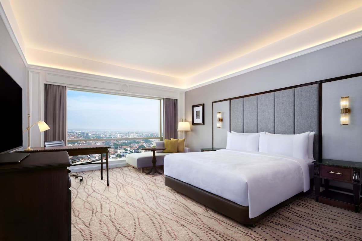 A hotel room with a large bed, a desk, a chair, a TV, and a view of the city.