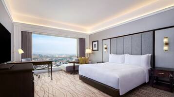 A hotel room with a large bed, a desk, a chair, a TV, and a view of the city.