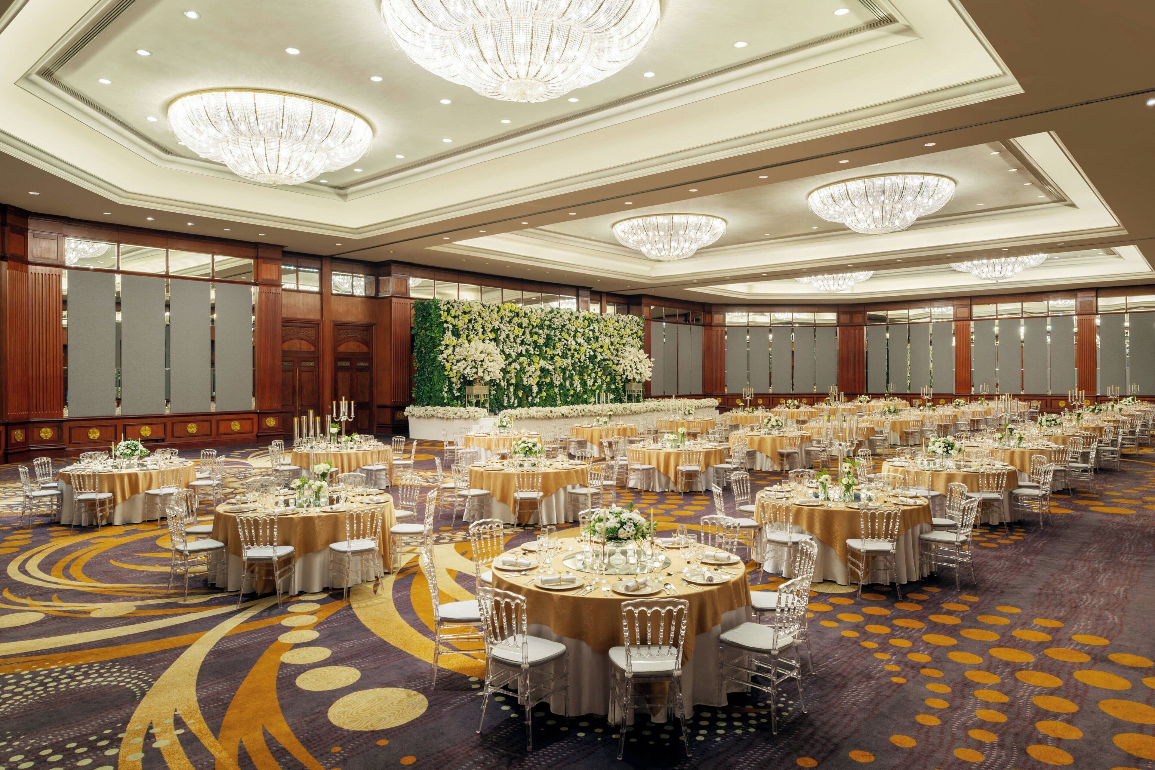 A large banquet hall with round tables, chandeliers, and a floral wall decoration.