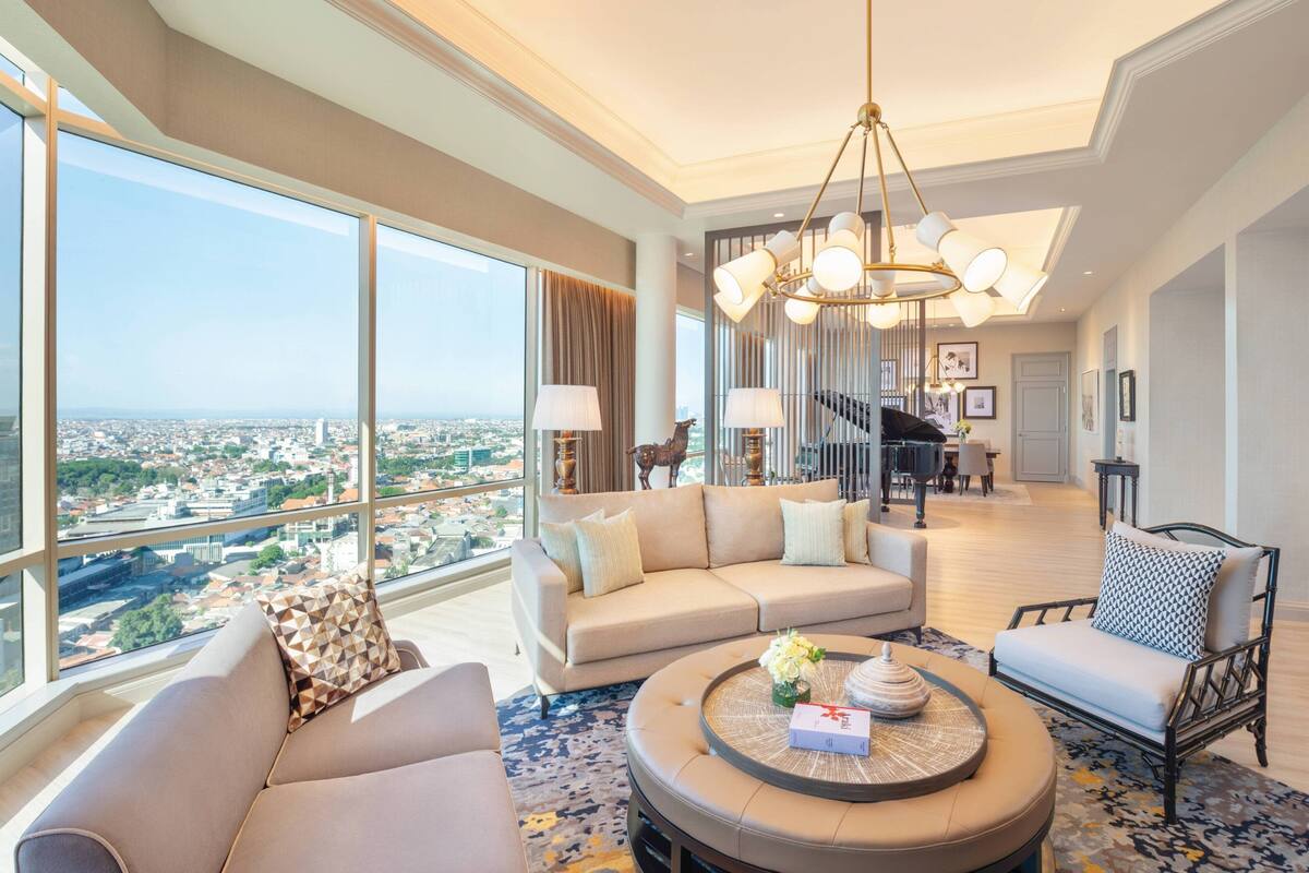 A spacious living room with a large window offering a city view, a round coffee table, and a modern chandelier.