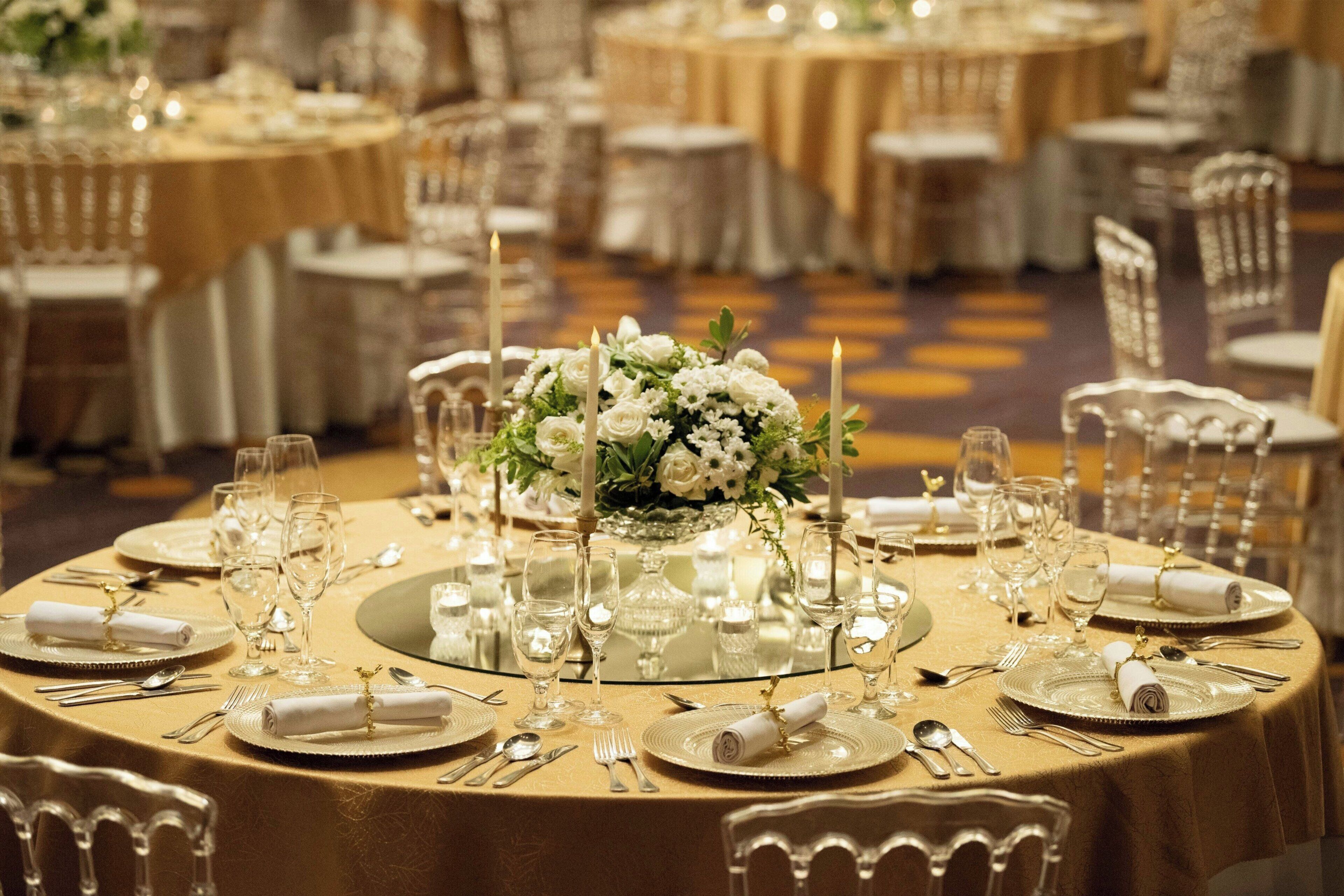 Un salón de banquetes con mesas redondas dispuestas para un evento formal, destacando manteles dorados, servilletas blancas y centros de mesa florales.