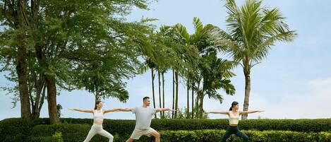 Tres personas practicando yoga al aire libre en un campo cubierto de césped, con árboles y un seto de fondo.