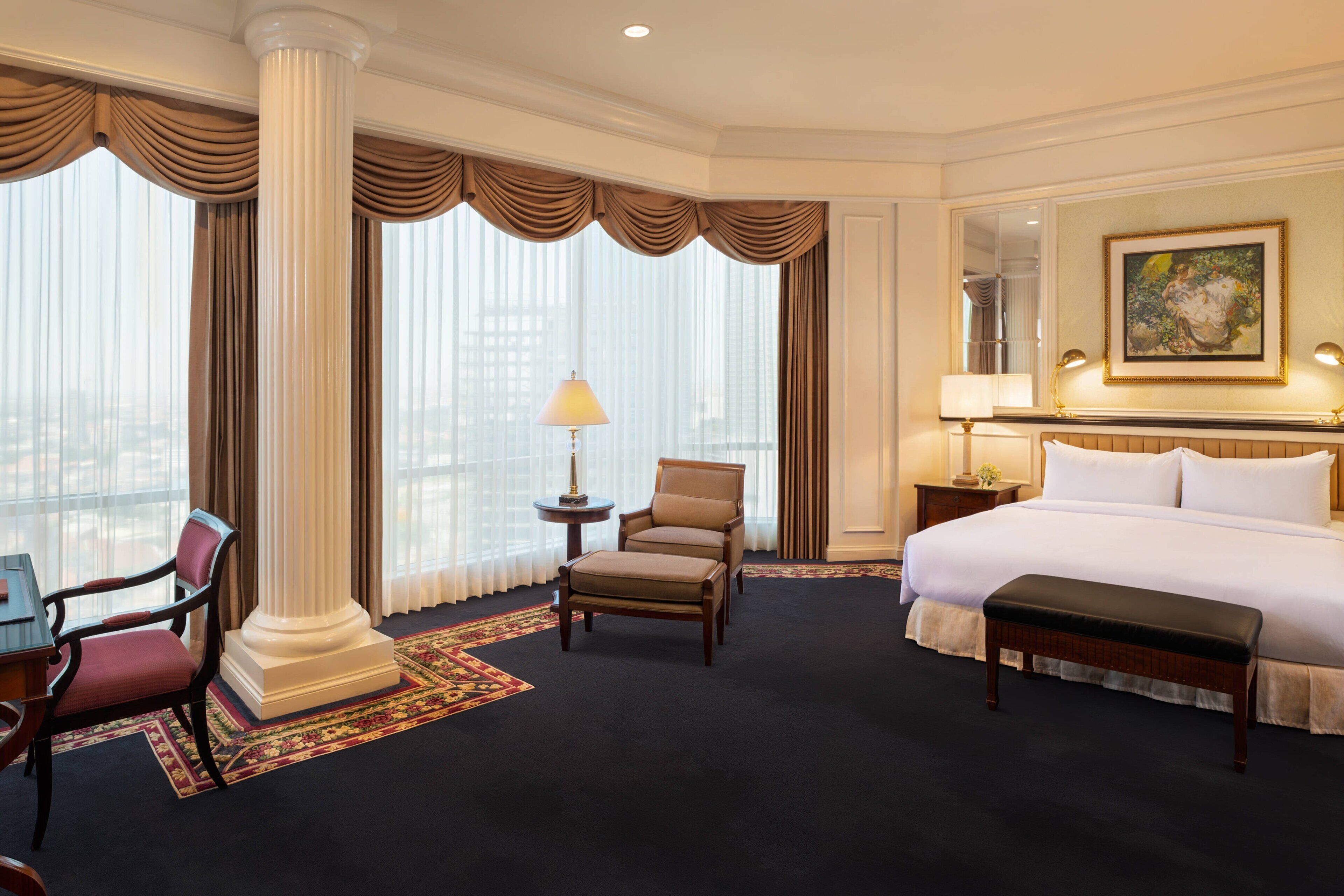 A hotel room with a large bed, a desk with a chair, a small table with a lamp, and a large window with curtains.