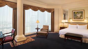 A hotel room with a large bed, a desk with a chair, a small table with a lamp, and a large window with curtains.