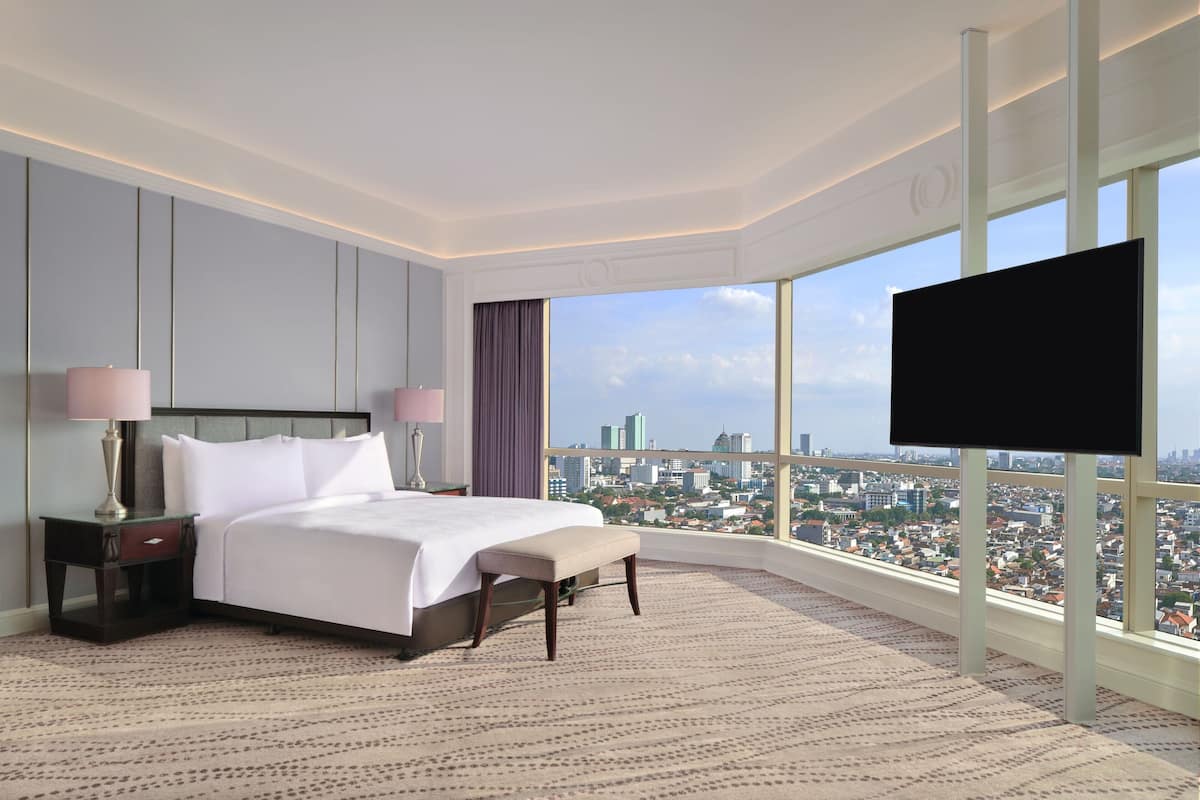A modern hotel room with a large bed, a flat-screen TV, and a city view through large windows.
