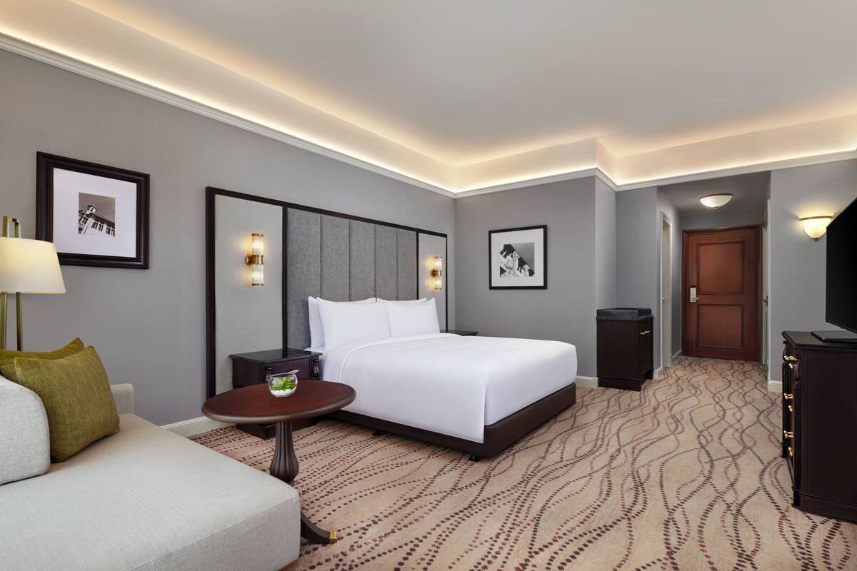 A hotel room with a large bed, a bedside table, a lamp, framed pictures on the wall, and a carpeted floor.