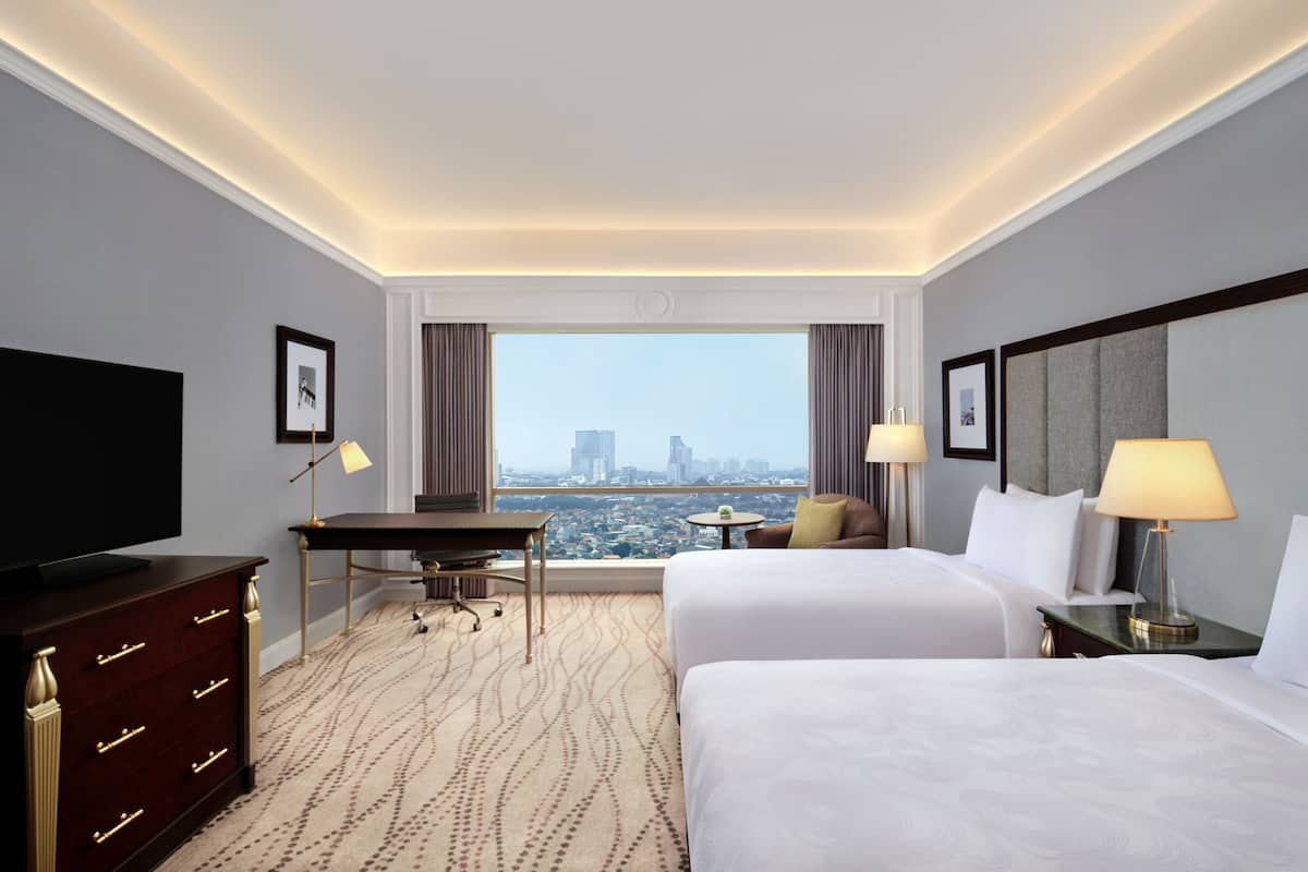 A hotel room with two beds, a desk, a television, and a view of a cityscape.
