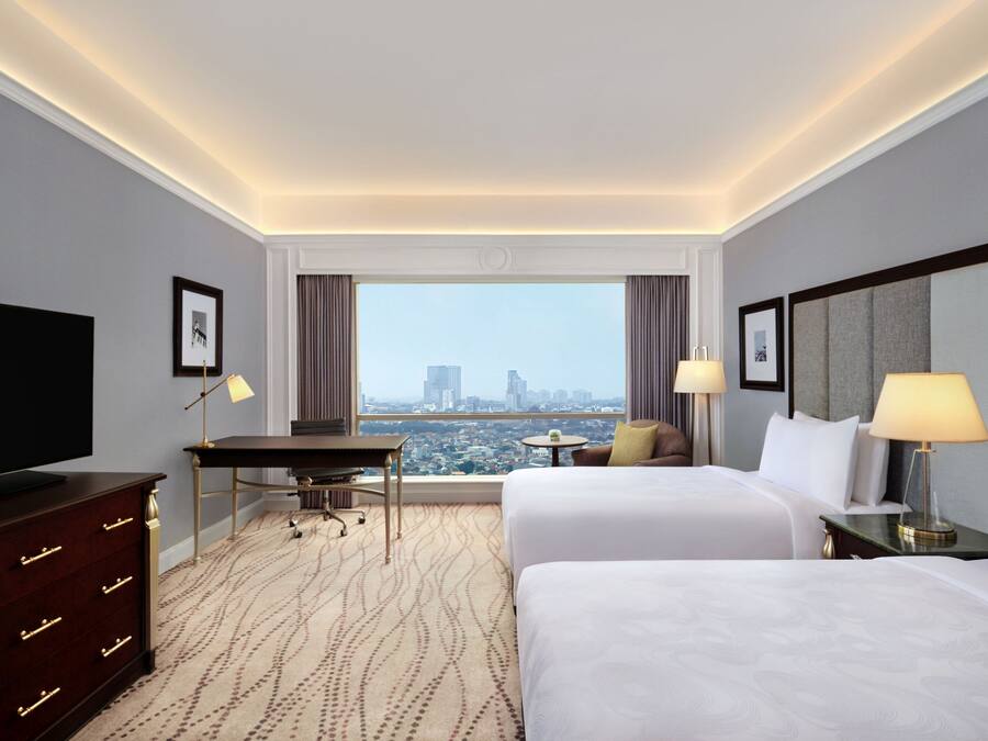A hotel room with two beds, a desk, a television, and a view of a cityscape.