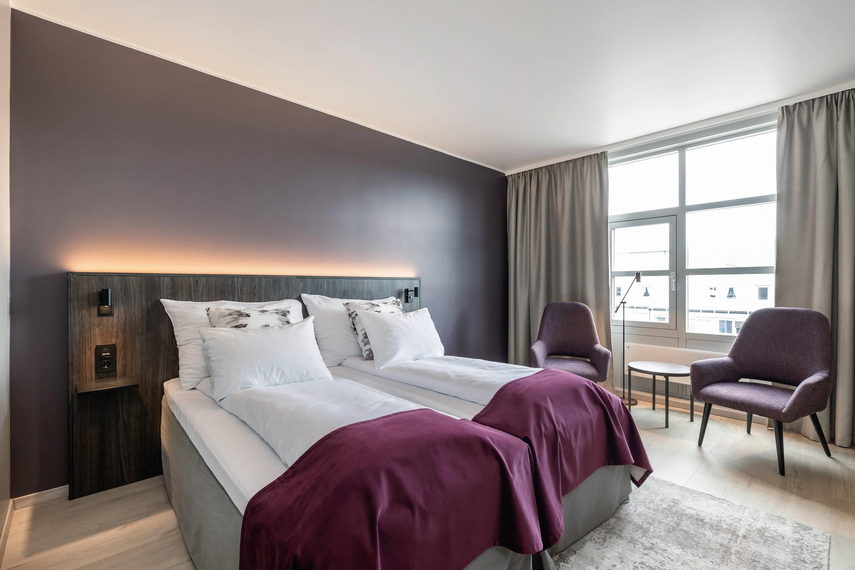 Et moderne hotelværelse med en stor seng, to sengeborde, en stol, et lille bord og et stort vindue med gardiner.