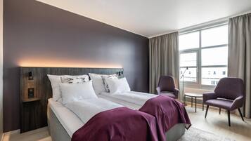 Et moderne hotelværelse med en stor seng, to sengeborde, en stol, et lille bord og et stort vindue med gardiner.