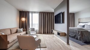 Una habitación de hotel moderna con sofá, sillón, televisor, cama y un ventanal grande.