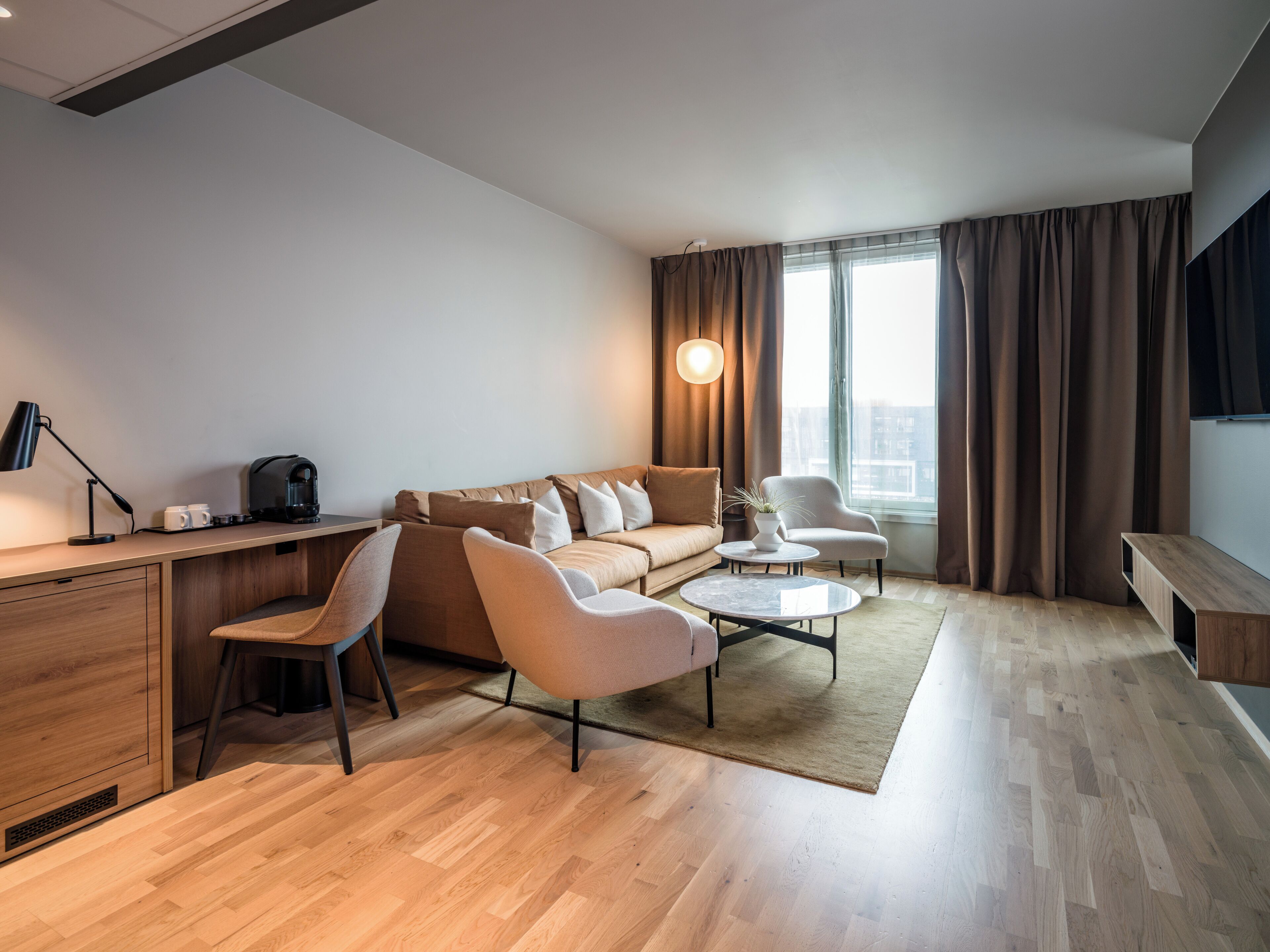 A modern hotel room with a sofa, armchairs, a coffee table, a desk with a lamp, and a large window with curtains.