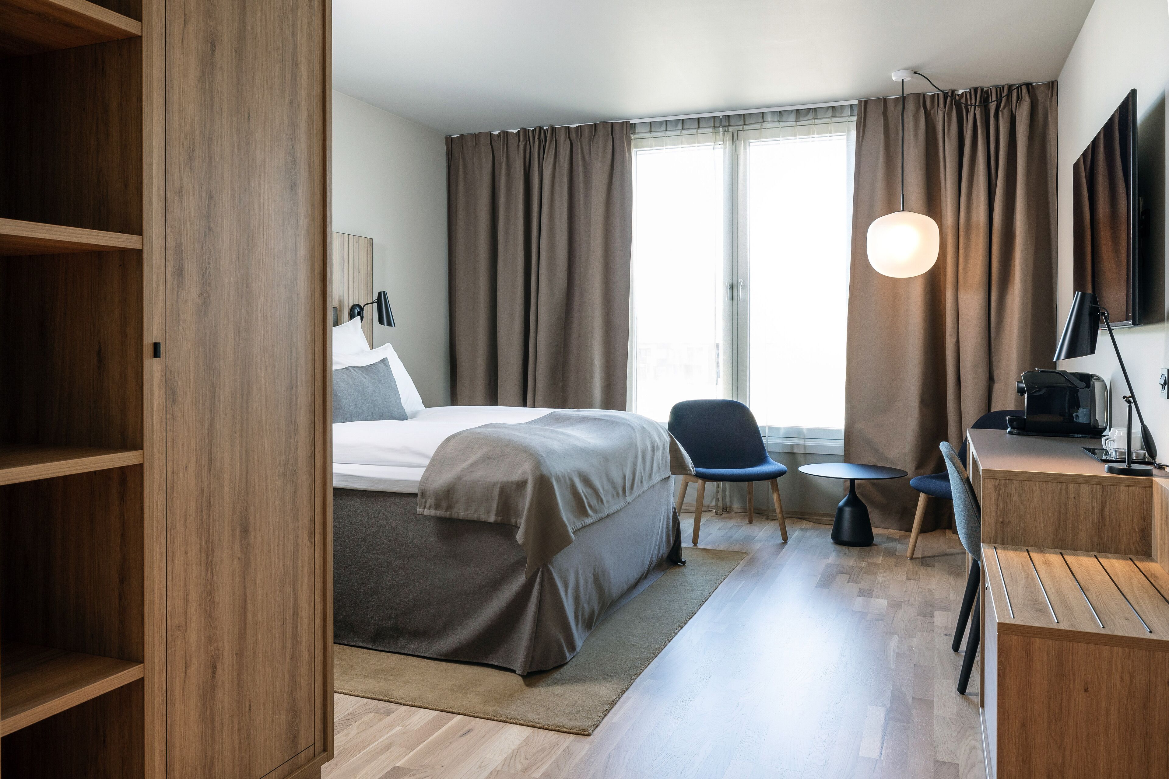 Habitación de hotel con cama, escritorio, silla, televisor y ventana con cortinas.