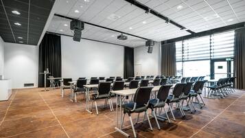 Un salón de conferencias con filas de sillas y mesas, un proyector y un ventanal con cortinas.