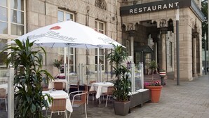 An outdoor seating area with tables and chairs, potted plants, and a large umbrella with a logo. - Wyndham Duisburger Hof (Duisburg)