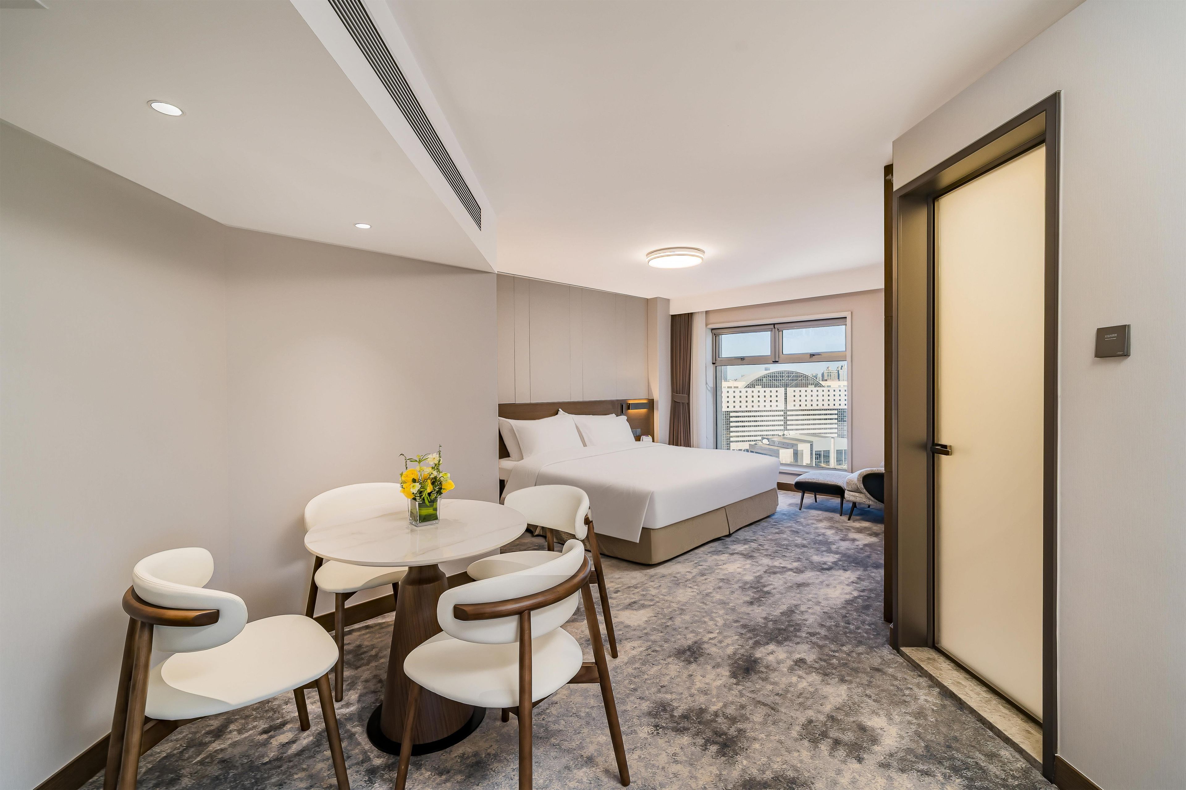 A hotel room with a bed, a small round table with chairs, a vase of flowers, and a view of the city through the window.