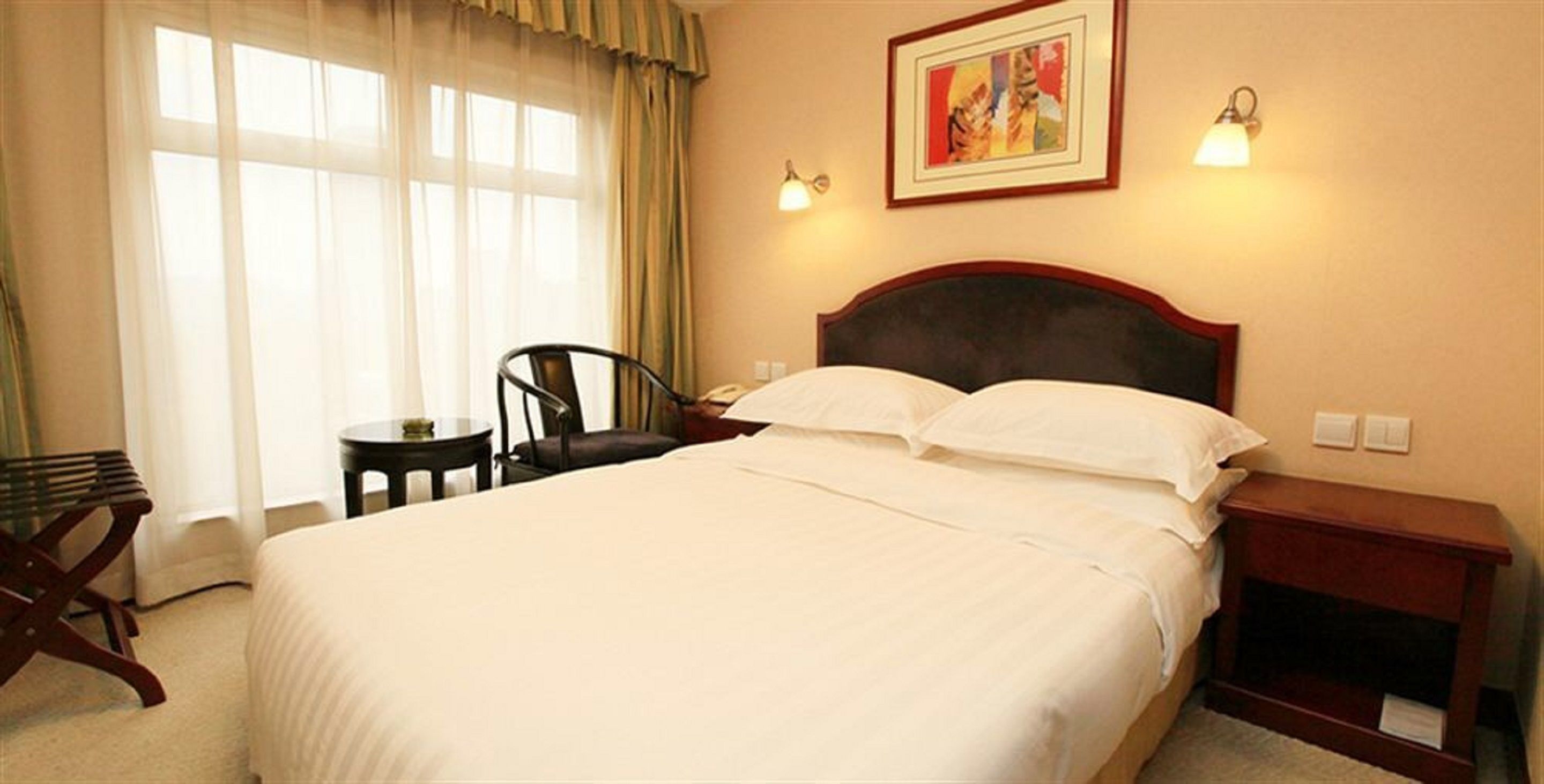 A hotel room with a large bed, a bedside table, a chair, a desk, and a painting on the wall.