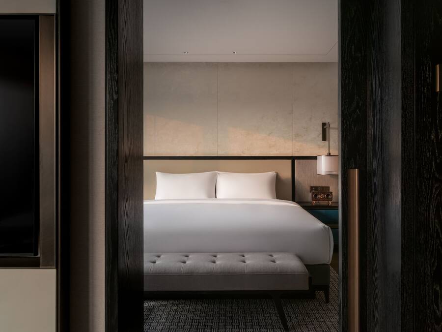 Una habitación de hotel moderna con una cama grande, un banco, una mesita de noche con lámpara y un televisor.