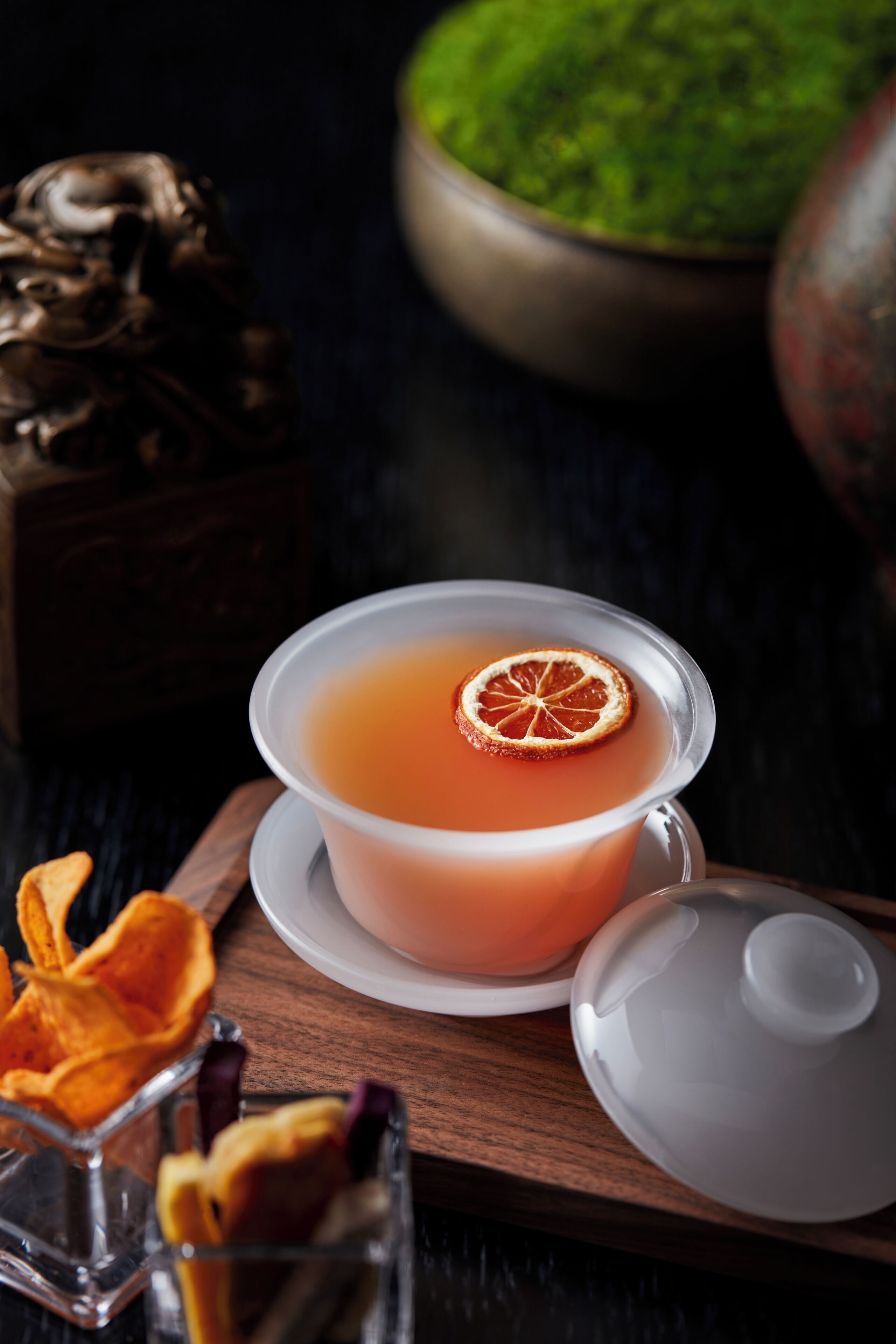 A clear glass cup with a lid, a slice of orange on the rim, and a wooden tray with a decorative item.