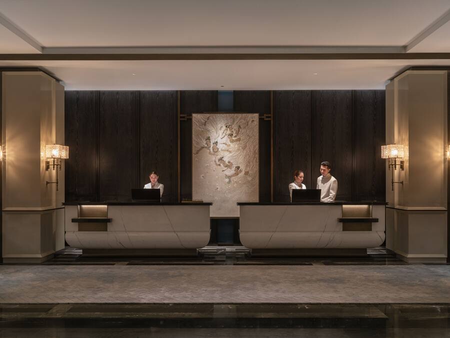Una zona de recepció amb dos recepcionistes darrere de taulells, una obra d'art abstracta de grans dimensions a la paret i làmpades penjades a la paret.