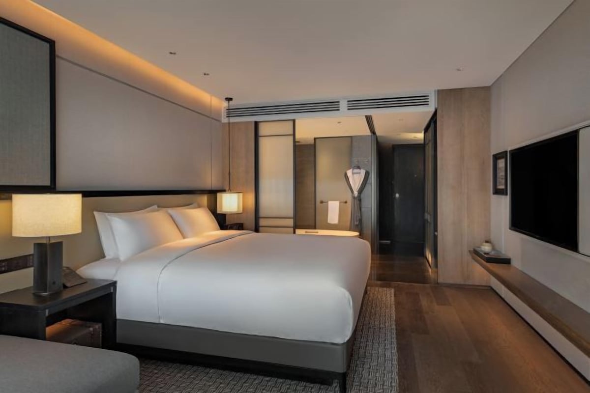 A modern hotel room with a large bed, bedside lamps, a flat-screen TV, and a view of the hallway.