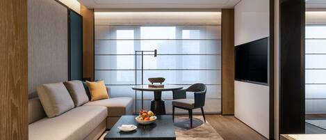 A modern living room with a sofa, coffee table, and chairs. A flat-screen TV is mounted on the wall.